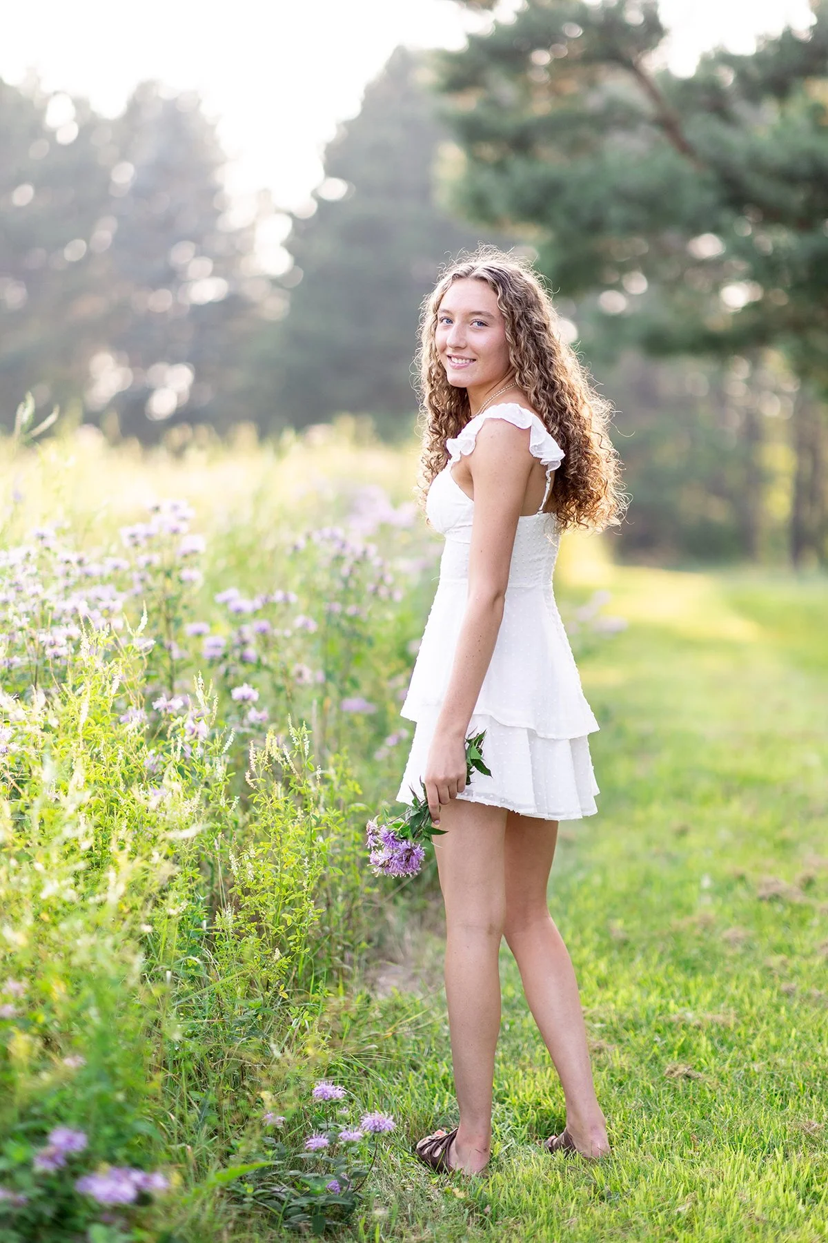 A young woman with curly hair in a white dress standing in a field of purple flowers, holding a bouquet of purple flowers, during golden hour with soft sunlight and trees in the background.