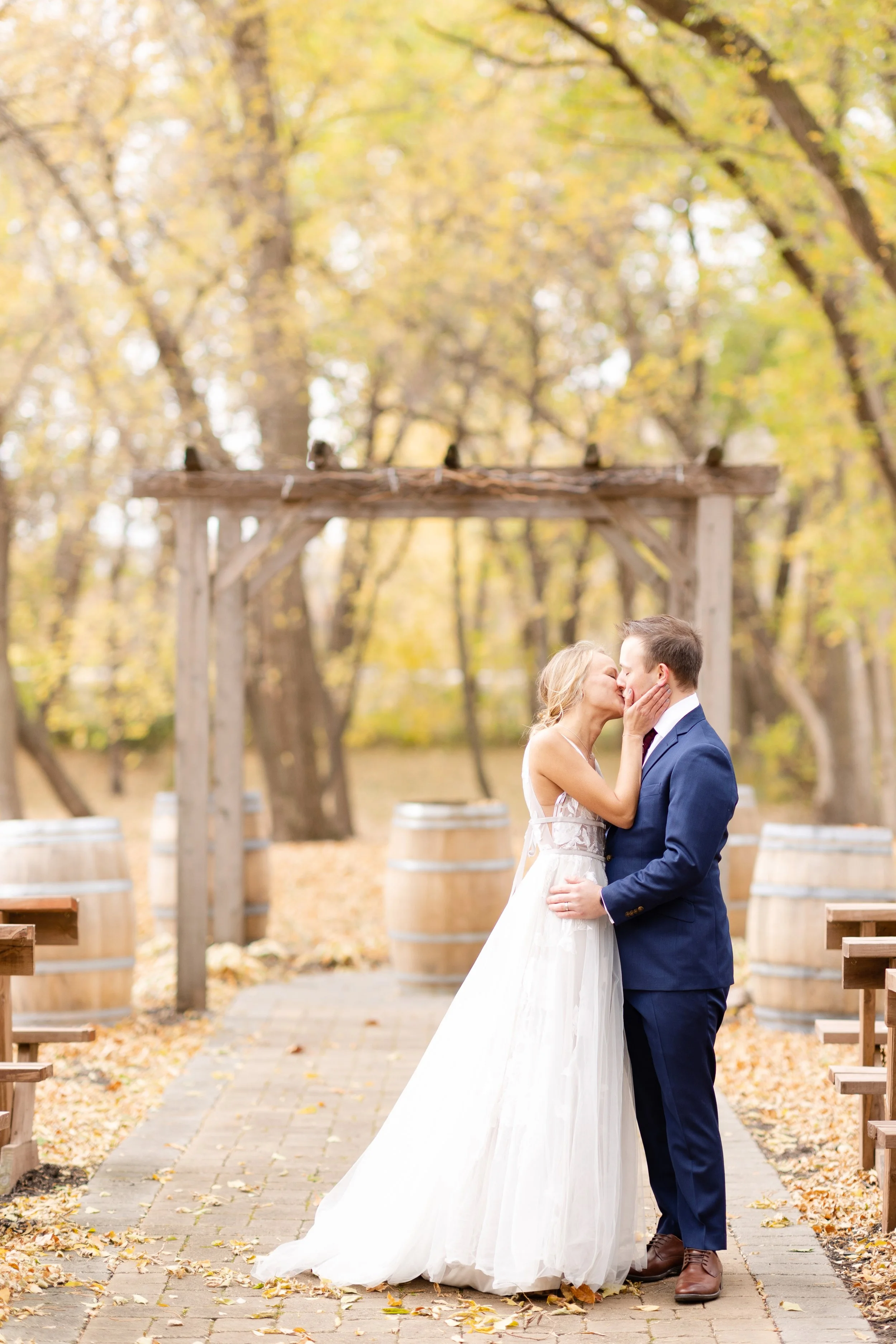 A bride and groom kissing during their wedding ceremony outdoors in a park with fall leaves and trees in the background.