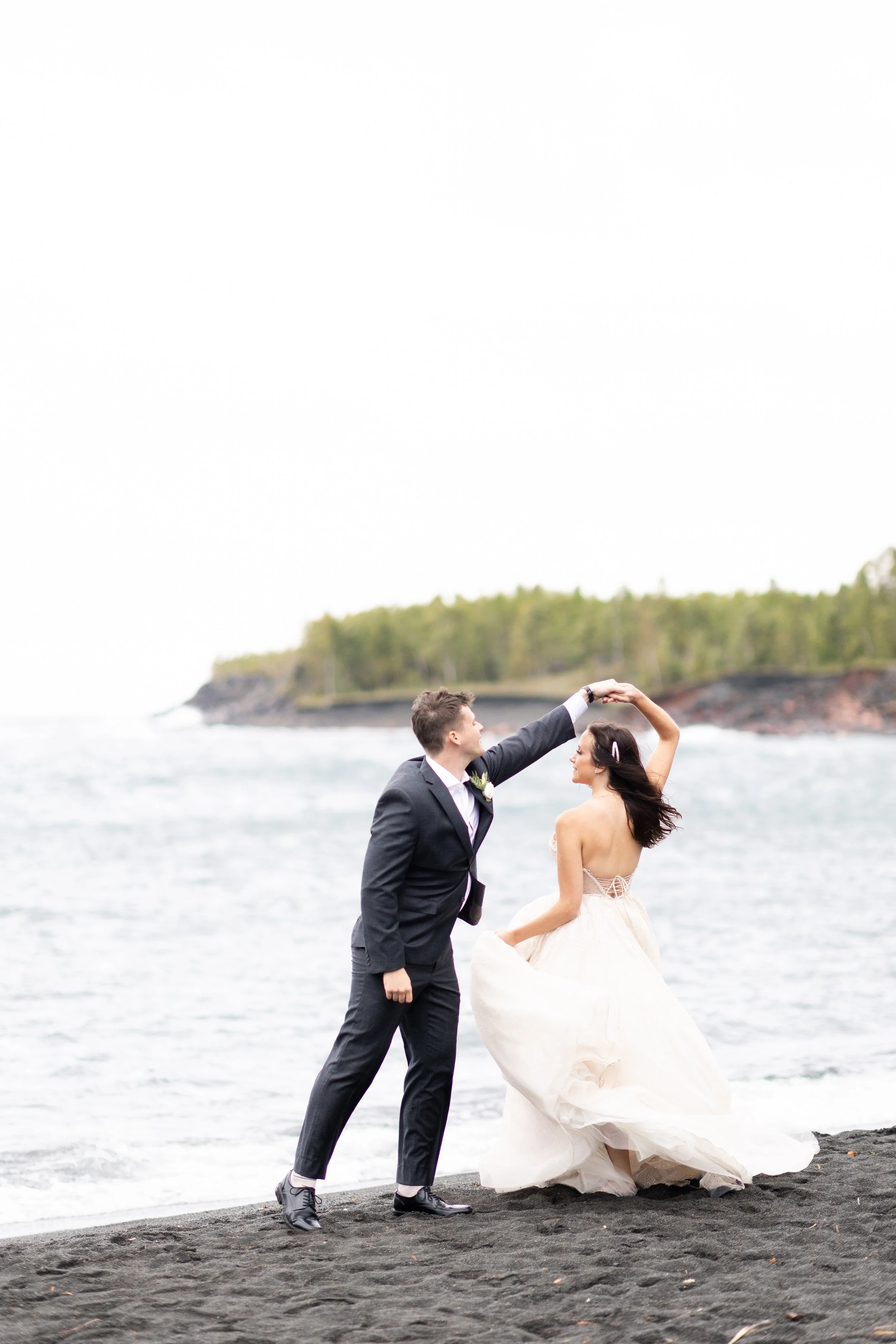 A newlywed couple dancing on a beach with black sand, with the ocean and forested coastline in the background.