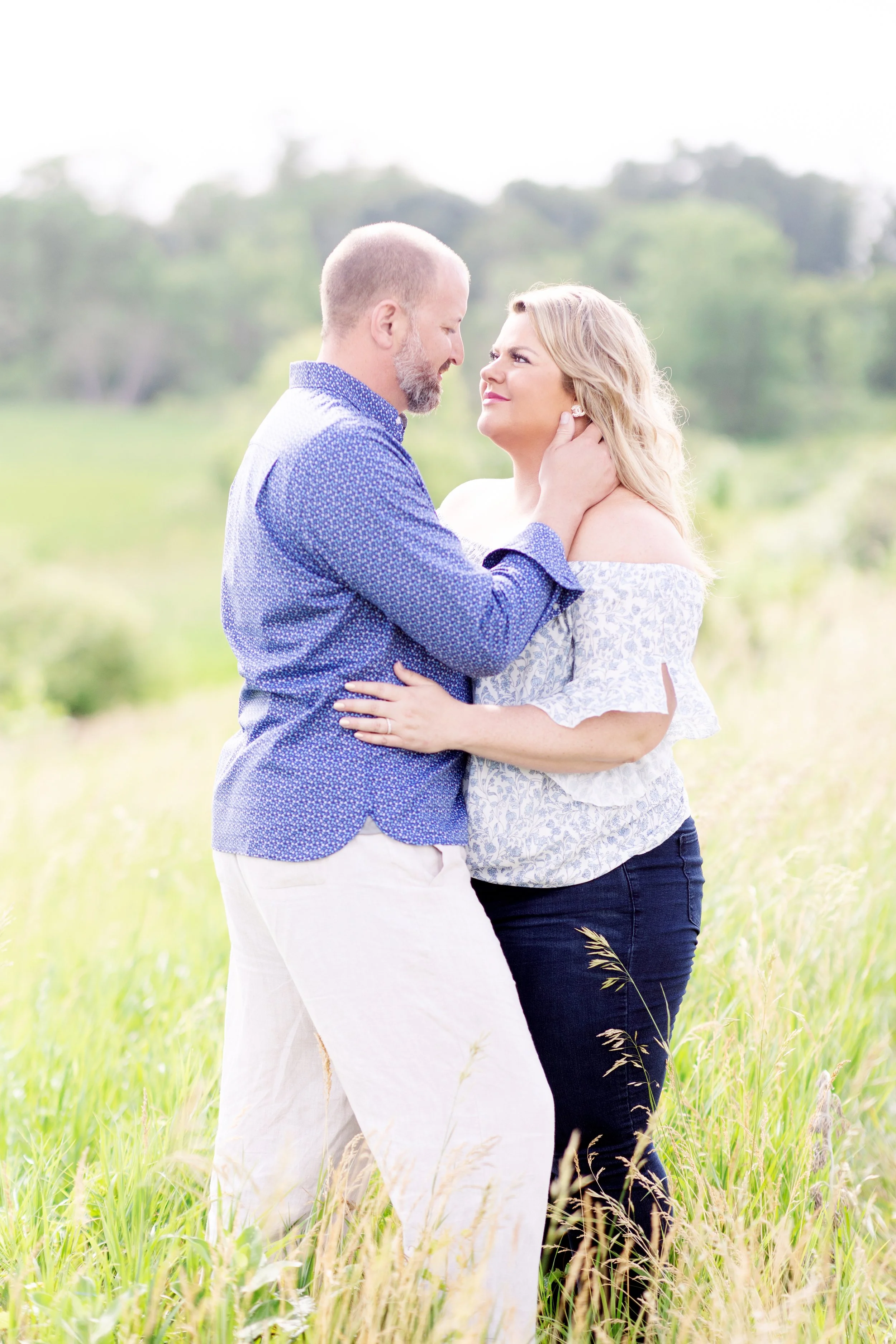A couple standing close together in a grassy field, gazing into each other's eyes with a blurred green landscape background.
