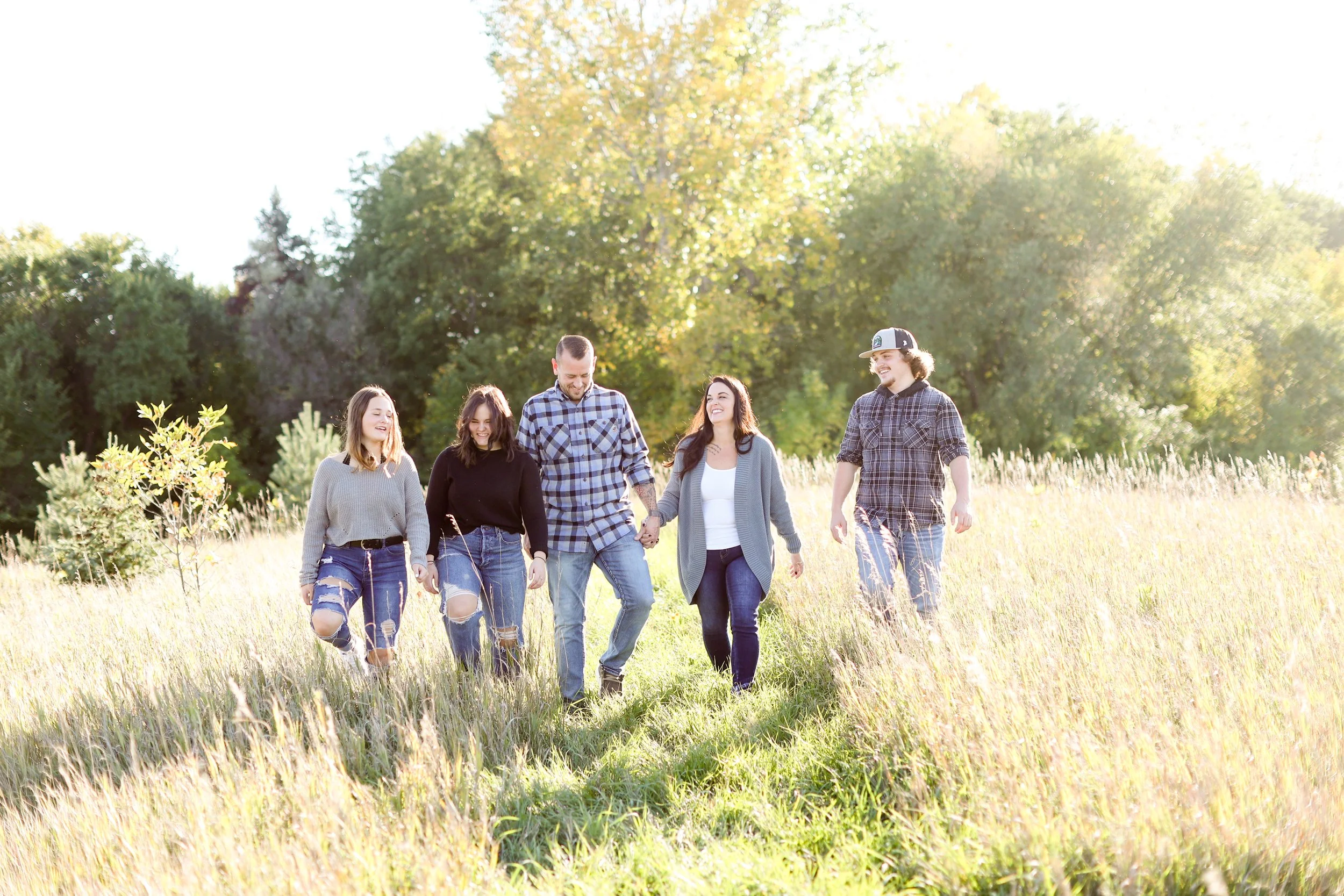 Five young adults walking hand in hand through a grassy field, smiling and enjoying sunny weather with trees in the background.
