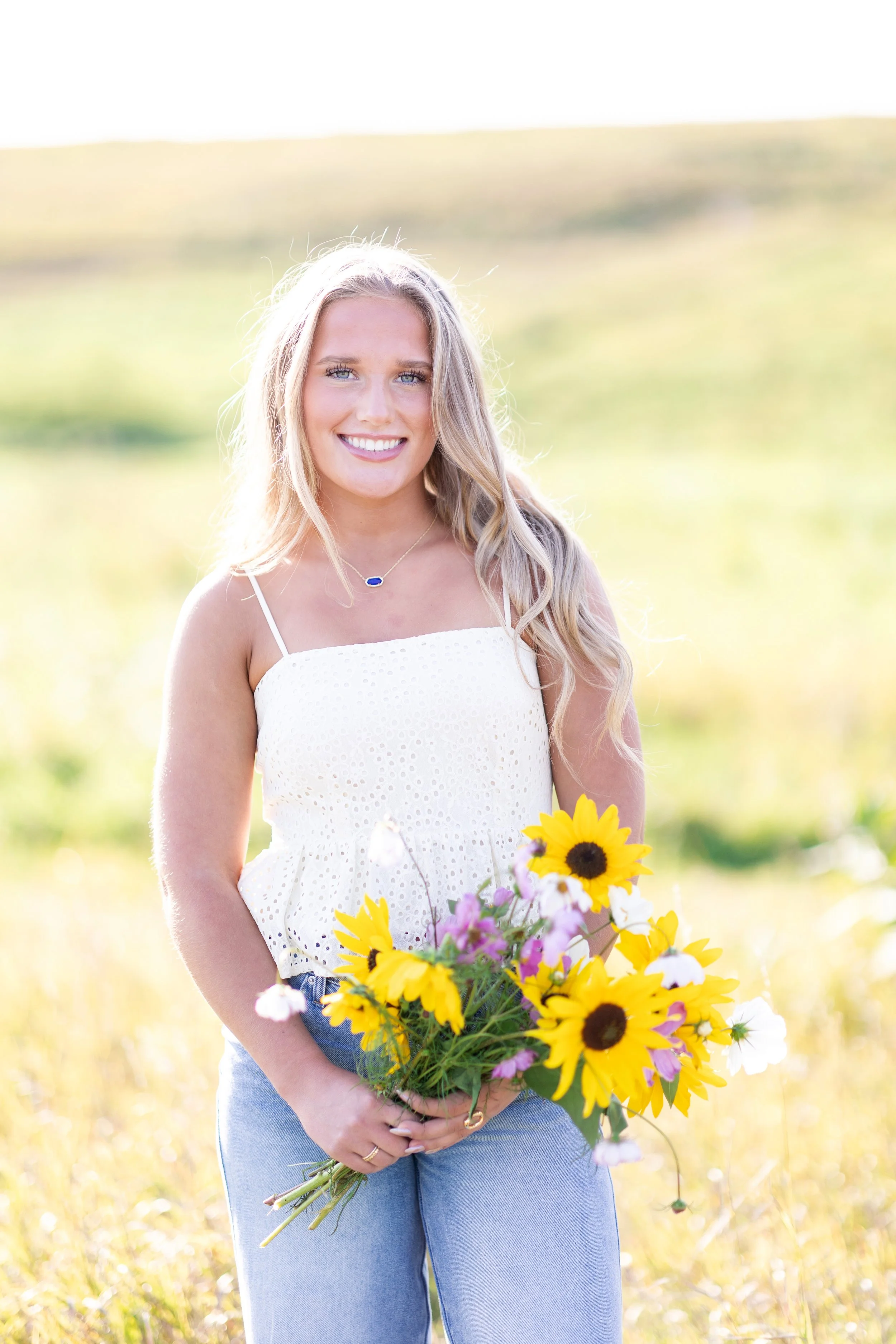 A smiling young woman with long blonde hair holding a colorful bouquet of sunflowers and daisies outdoors in a bright field.
