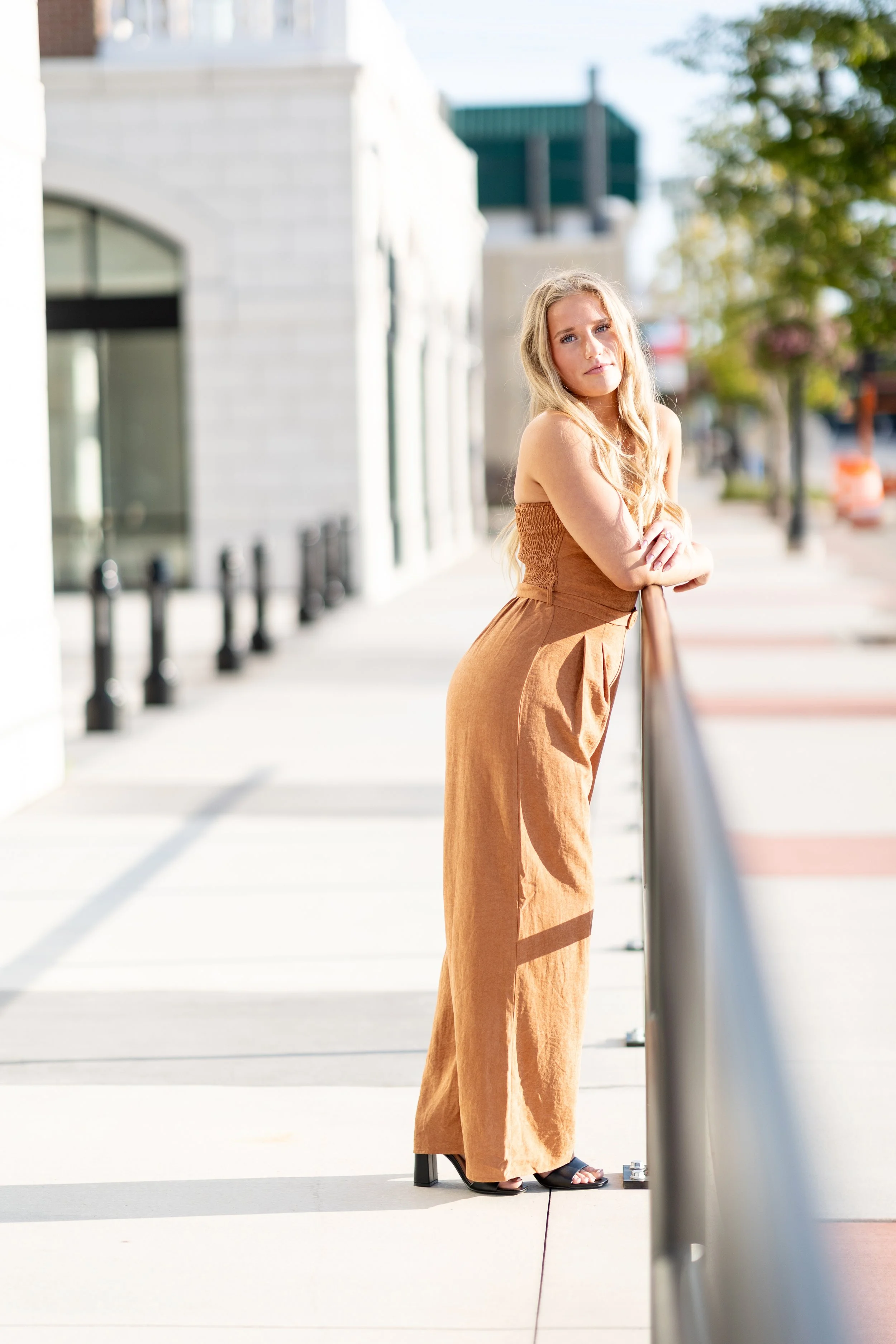 A woman leaning on a railing outdoors on a sunny day, wearing a strapless brown top and matching wide-leg pants with black sandals.