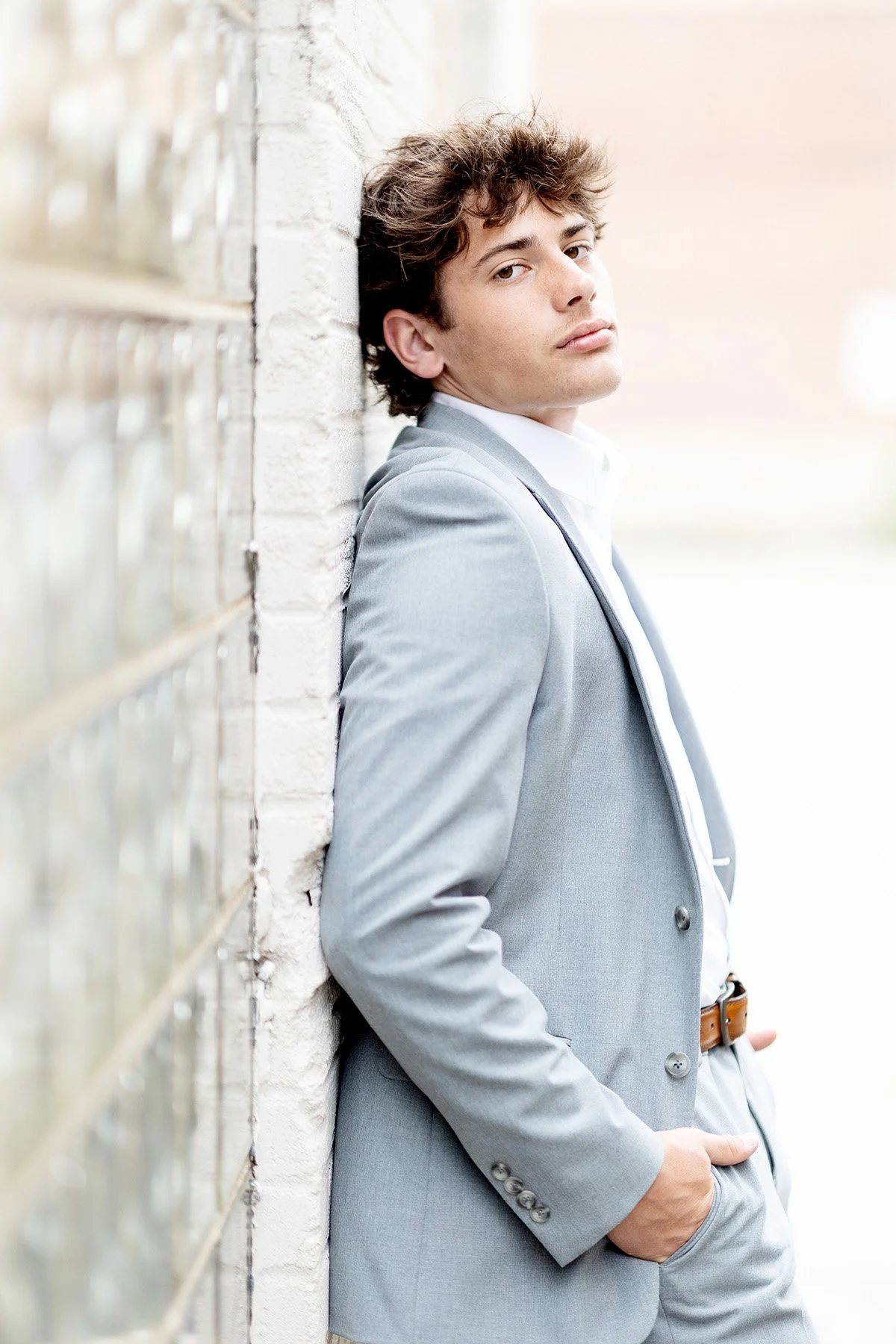 Young man in a gray suit leaning against a white brick wall, looking at the camera with a neutral expression.