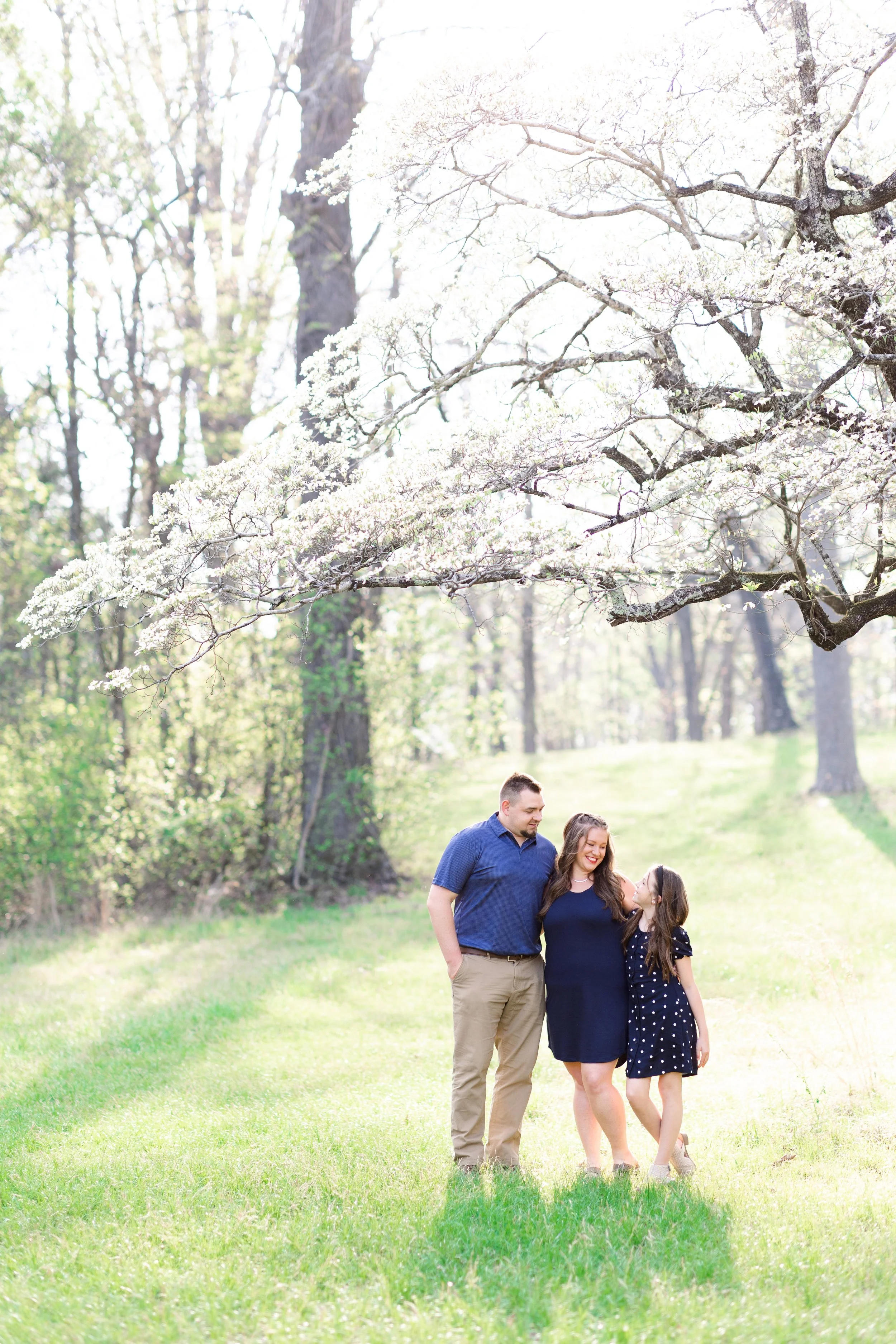 A family of three standing on a lush green lawn in a park during spring, with blooming trees and a bright sky overhead. The family includes a man, a woman, and a young girl, all smiling and looking at each other.