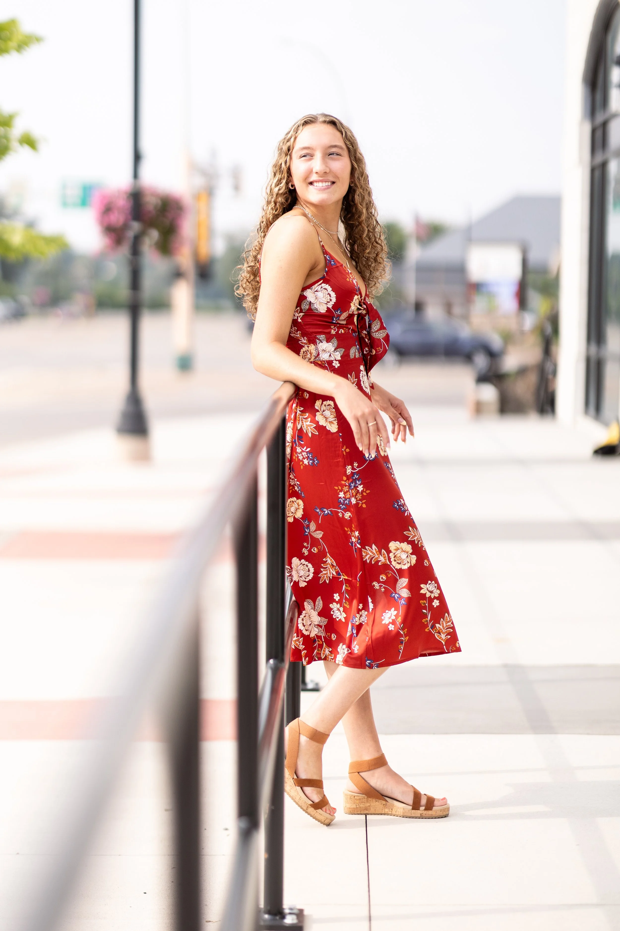 A young woman with curly hair smiling, wearing a red floral dress and tan wedge sandals, standing outdoors with a street and buildings in the background.
