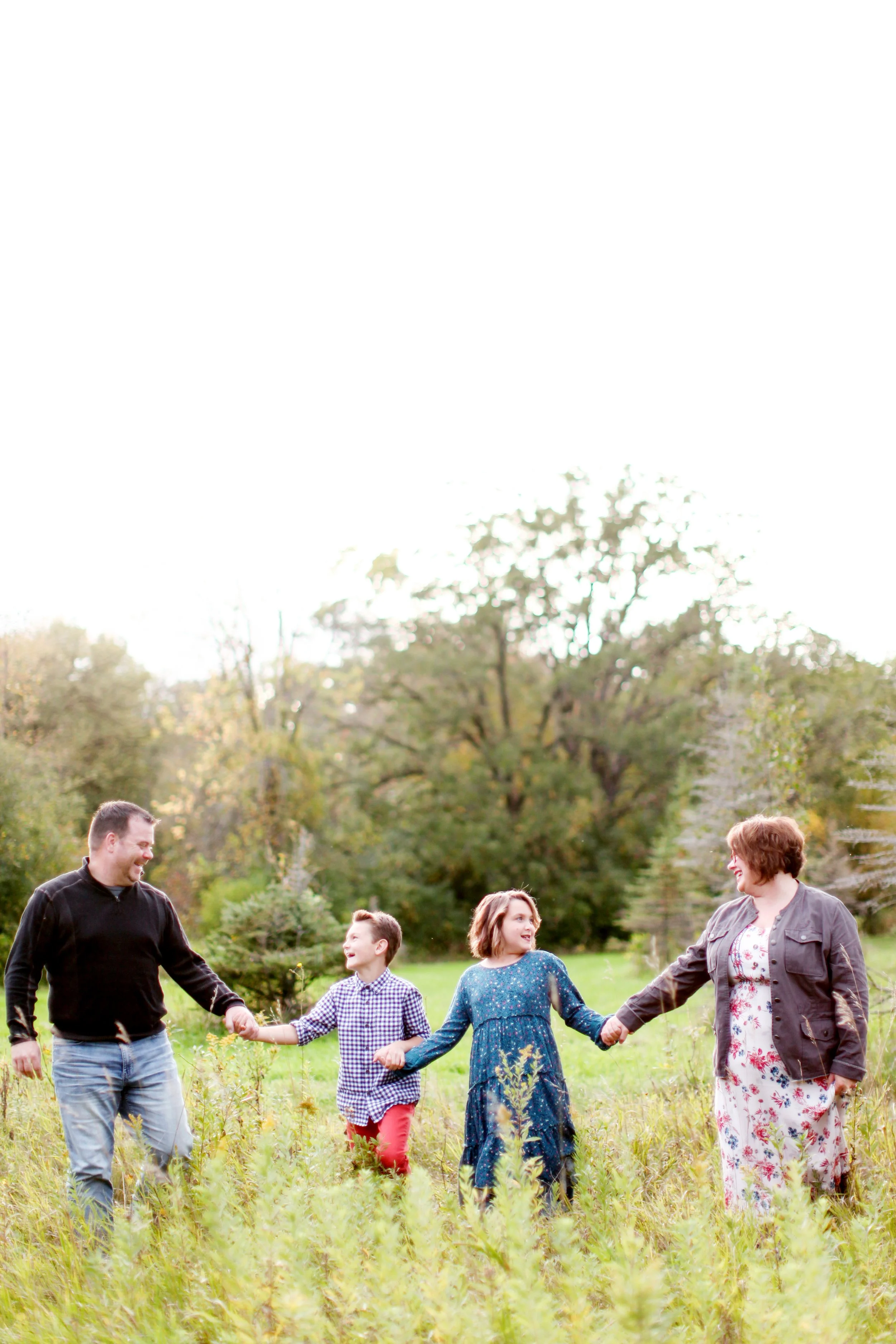 A family of five, holding hands and walking in a grassy field with trees in the background on a cloudy day.