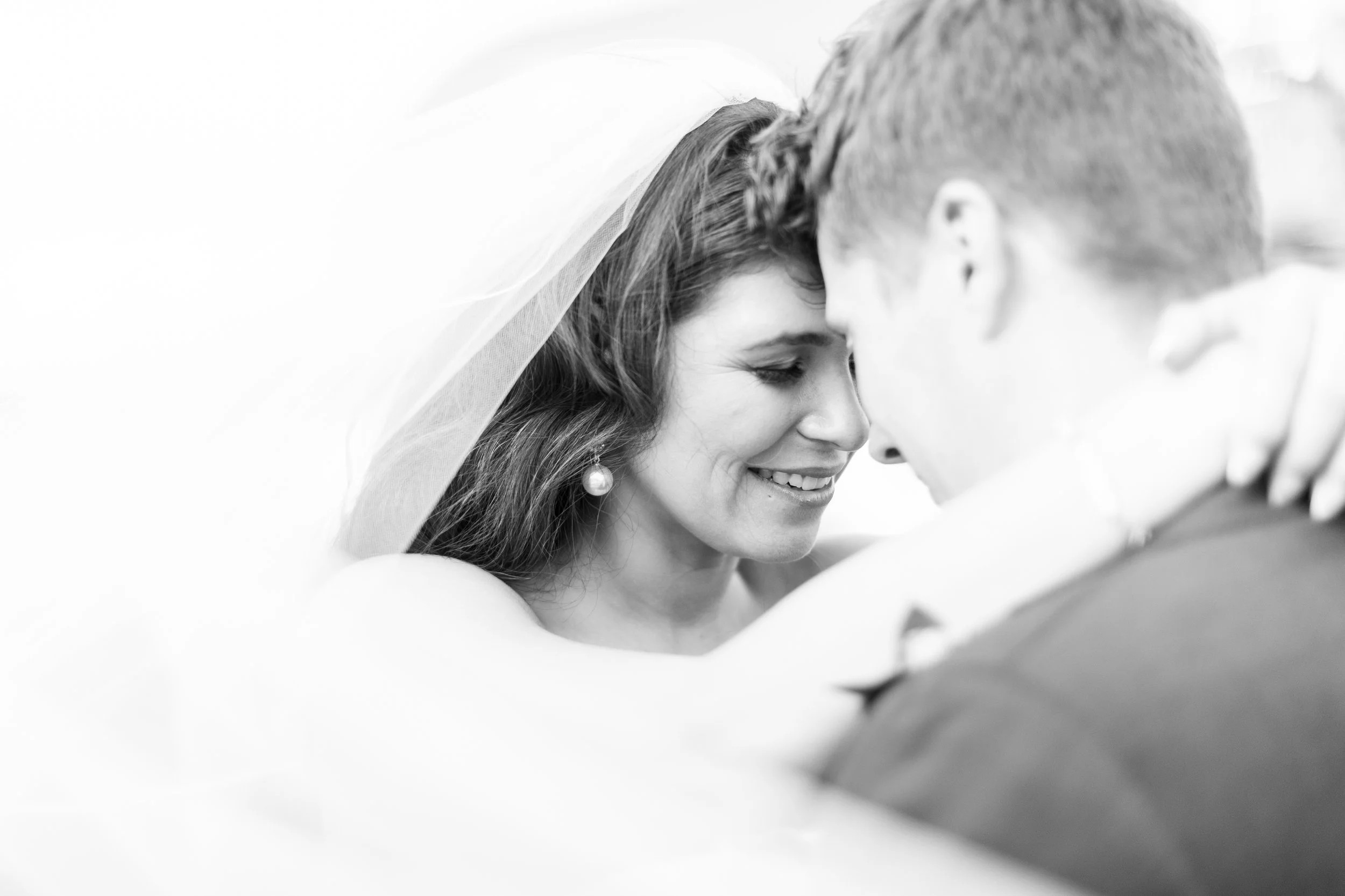 A black and white photograph of a woman with a veil leaning close to a man. They appear to be sharing an intimate moment, likely during a wedding.