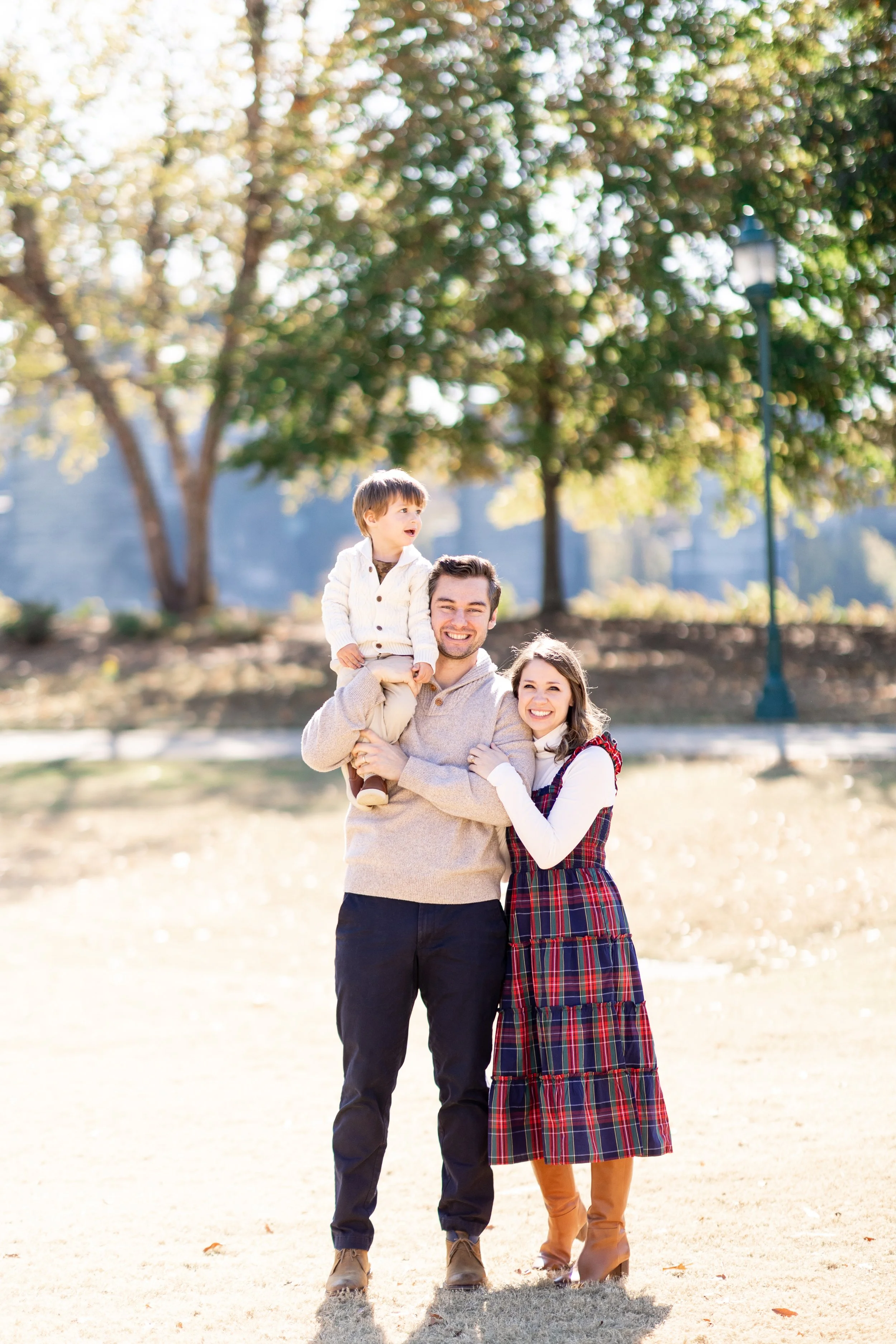 A family of three, a man, woman, and child, walking together outdoors in a park during daytime. The man is carrying the child on his shoulders, and everyone is smiling. The background features trees, a lamppost, and clear weather.