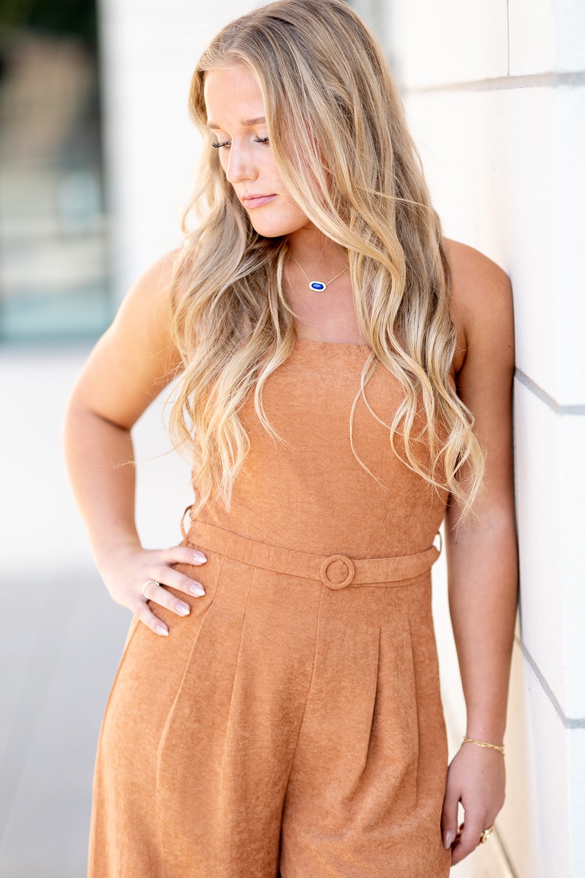 A woman with long, wavy blonde hair wearing a rust-colored sleeveless dress and standing against a white wall, looking down with her hand on her hip.