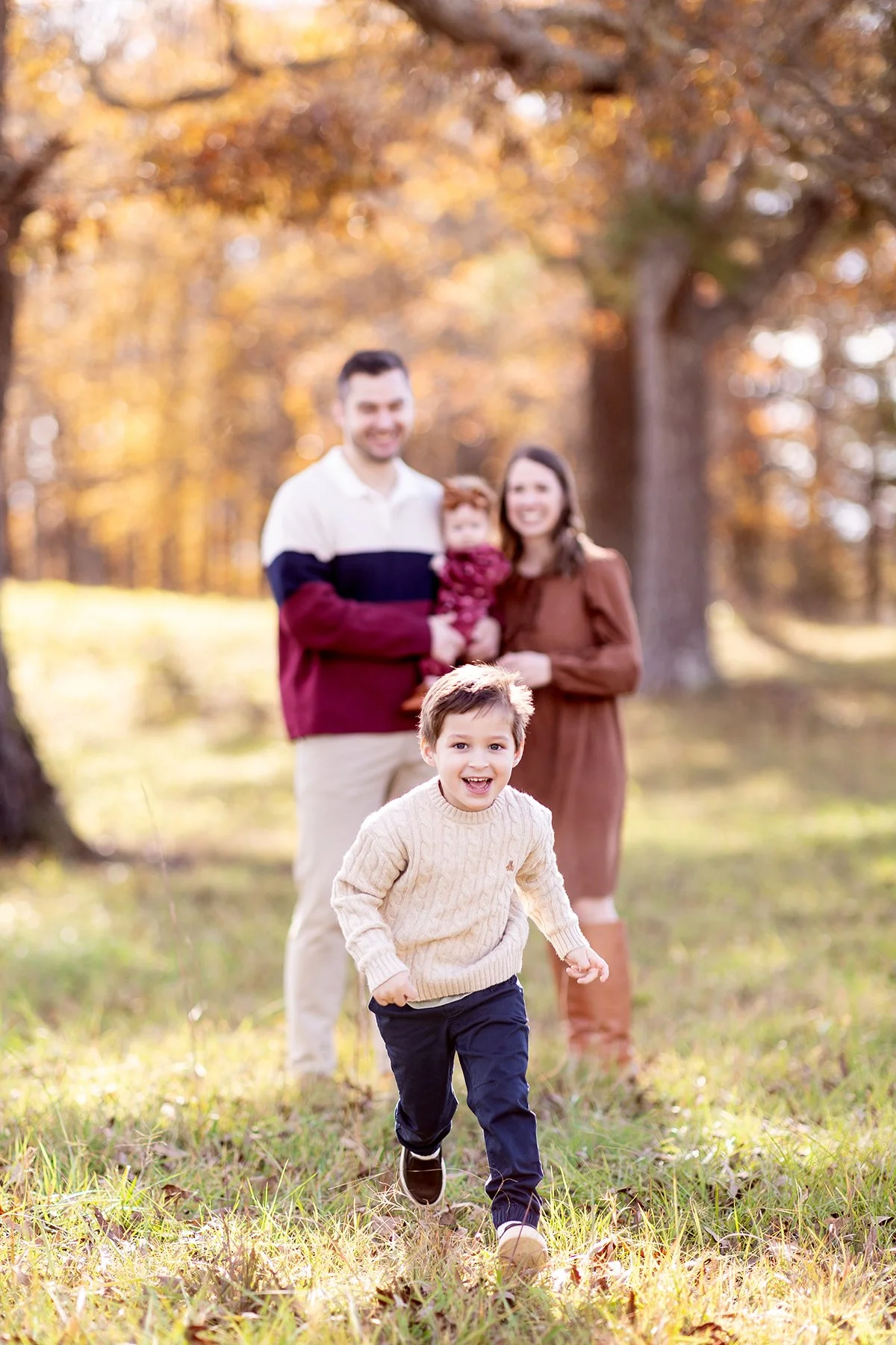 A young boy running and smiling in the foreground while a family of three stands in the background outdoors in a park with autumn trees.