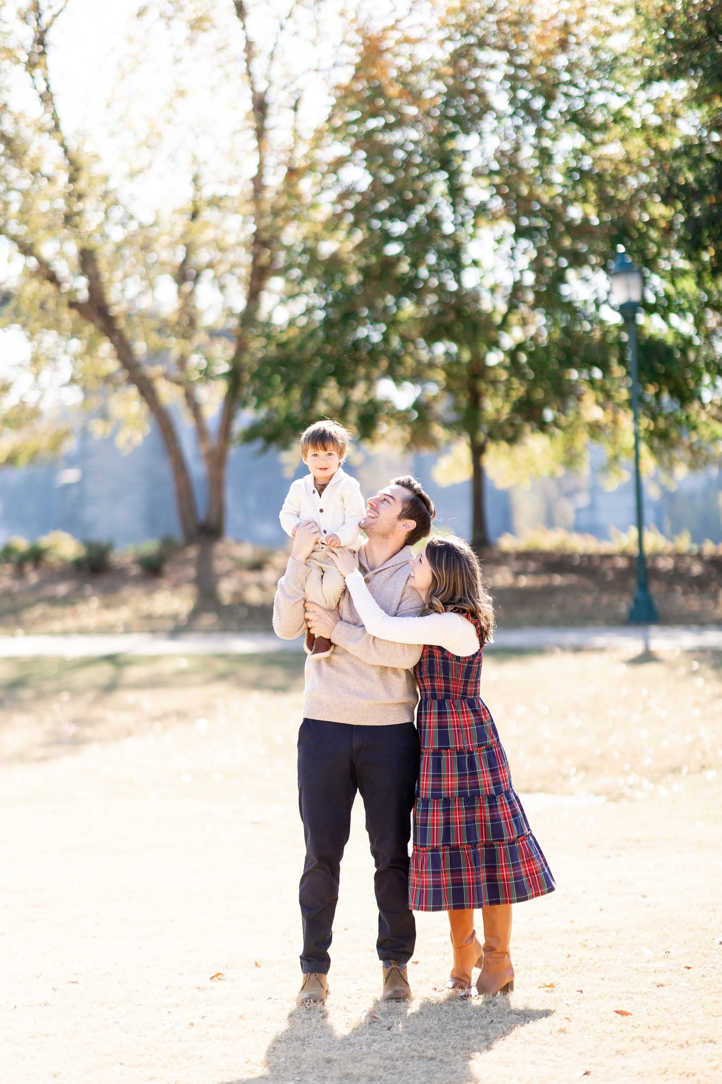A family outdoors in a park, with a man holding a young boy on his shoulders and a woman beside them. They are smiling and enjoying a sunny day with trees in the background.