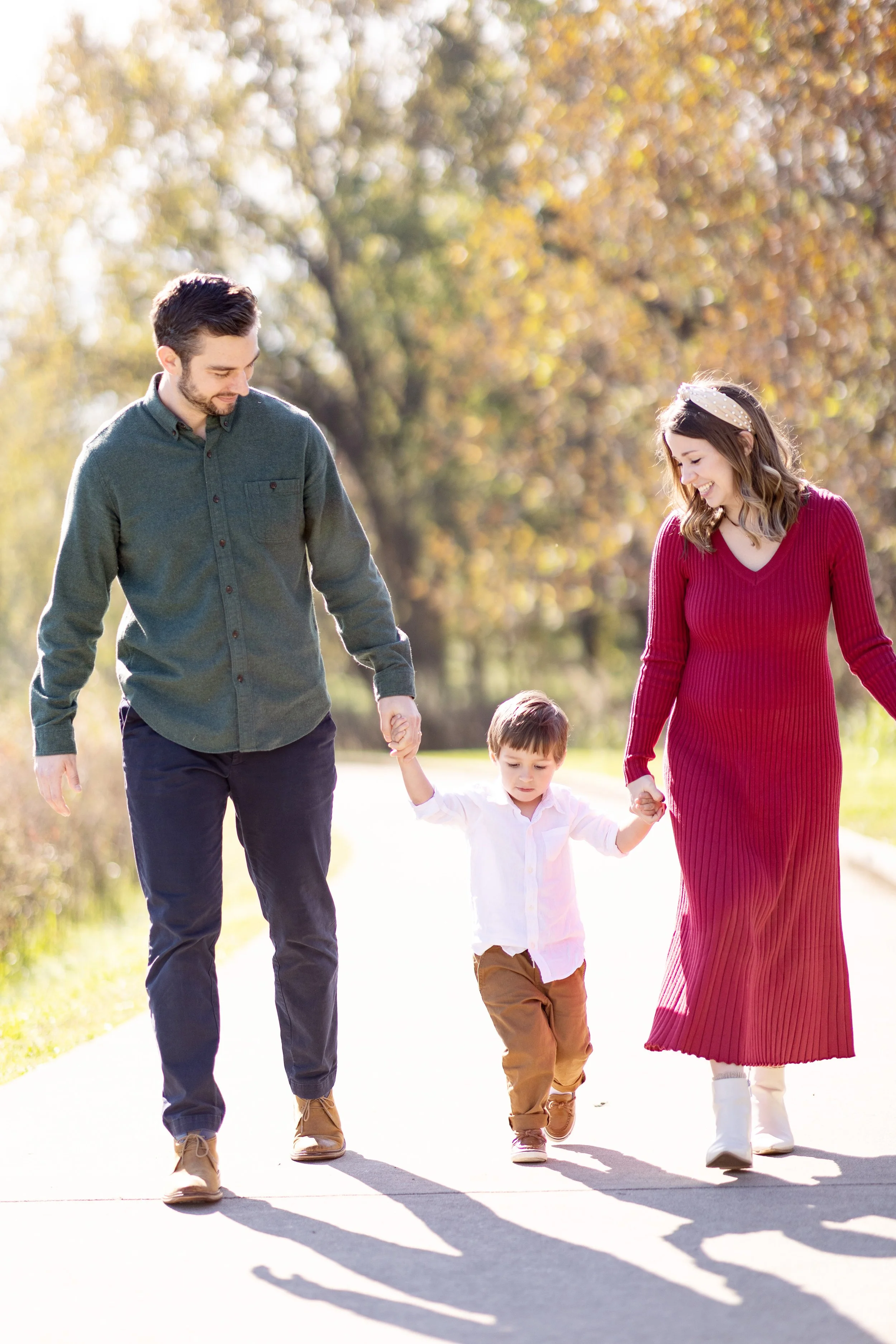 A family of four, including a father, a mother, and two children, walking outdoors during autumn. The father wears a dark green shirt and jeans, the mother wears a long red dress with a polka dot headband, the son wears a white shirt and tan pants, a