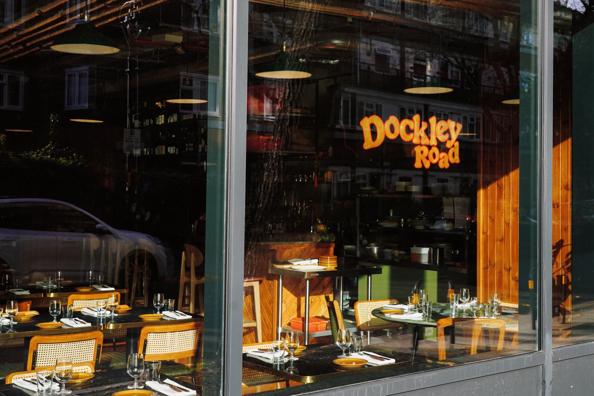 View of a restaurant interior through a large glass window, with tables set for dining, and a neon sign on the window that reads 'Dockley Road'.