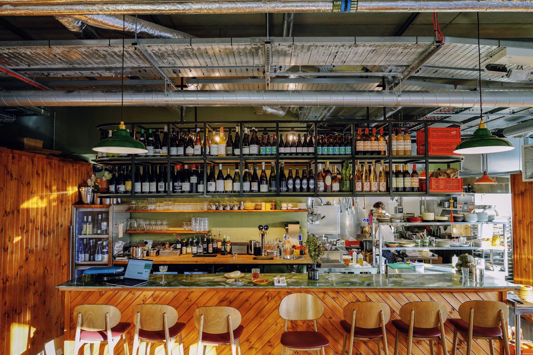 A restaurant interior with a bar area, shelves full of wine bottles, a green pendant light, and a kitchen in the background. Wooden wall paneling and chairs are visible in the foreground.