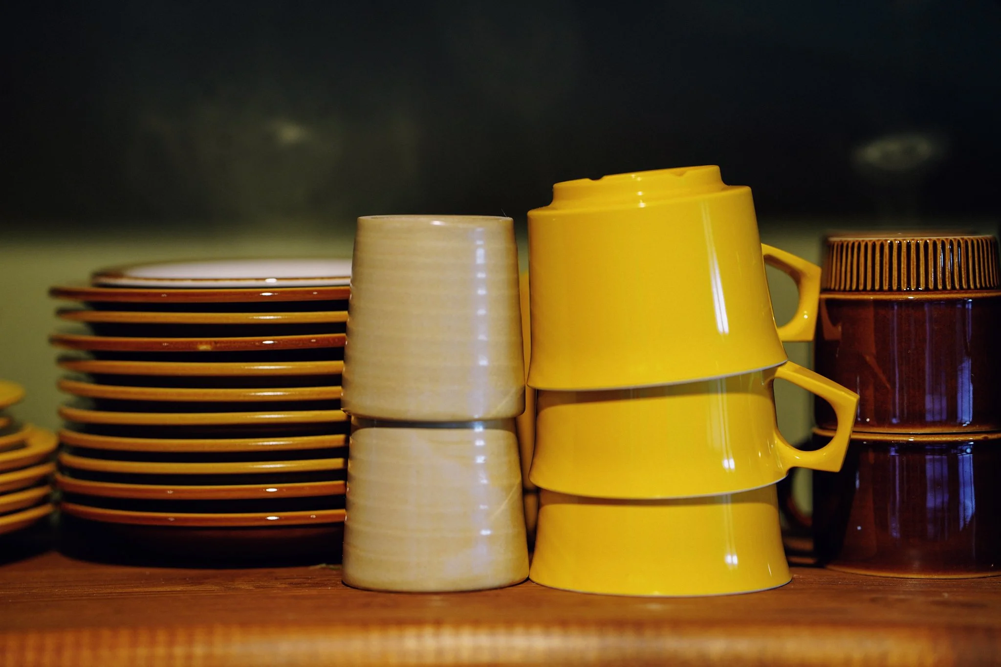 Stack of orange and brown plates on the left, beige and yellow mugs in the center, and a brown cup on the right, all placed on a wooden surface against a dark background.