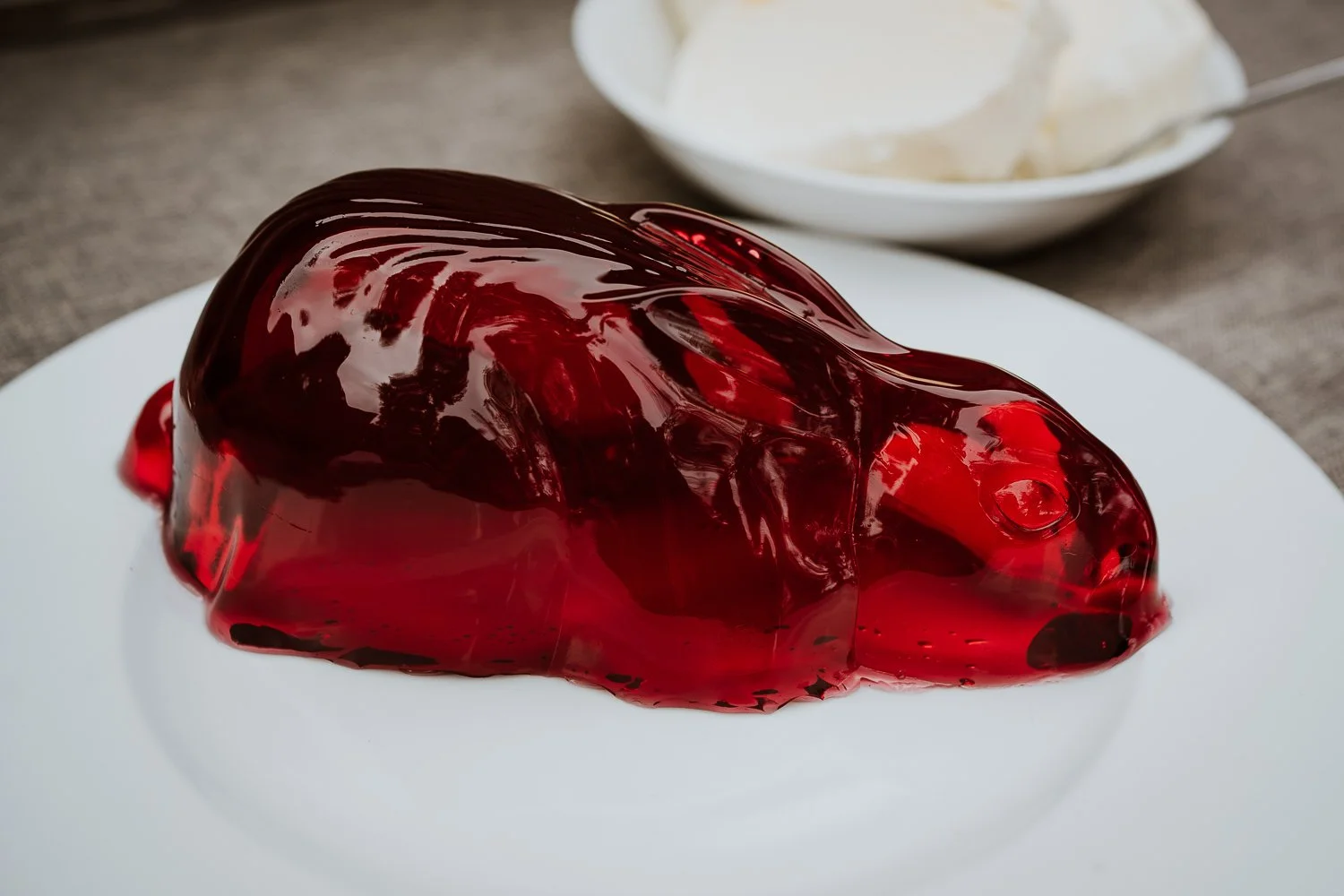 A glossy, red, transparent jelly-like dessert on a white plate with a bowl of white cream in the background.