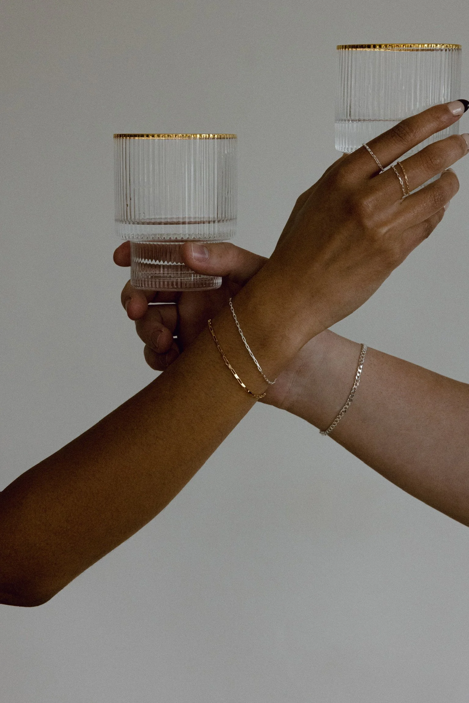 Two hands holding clear glass tumblers with gold rims against a plain background.