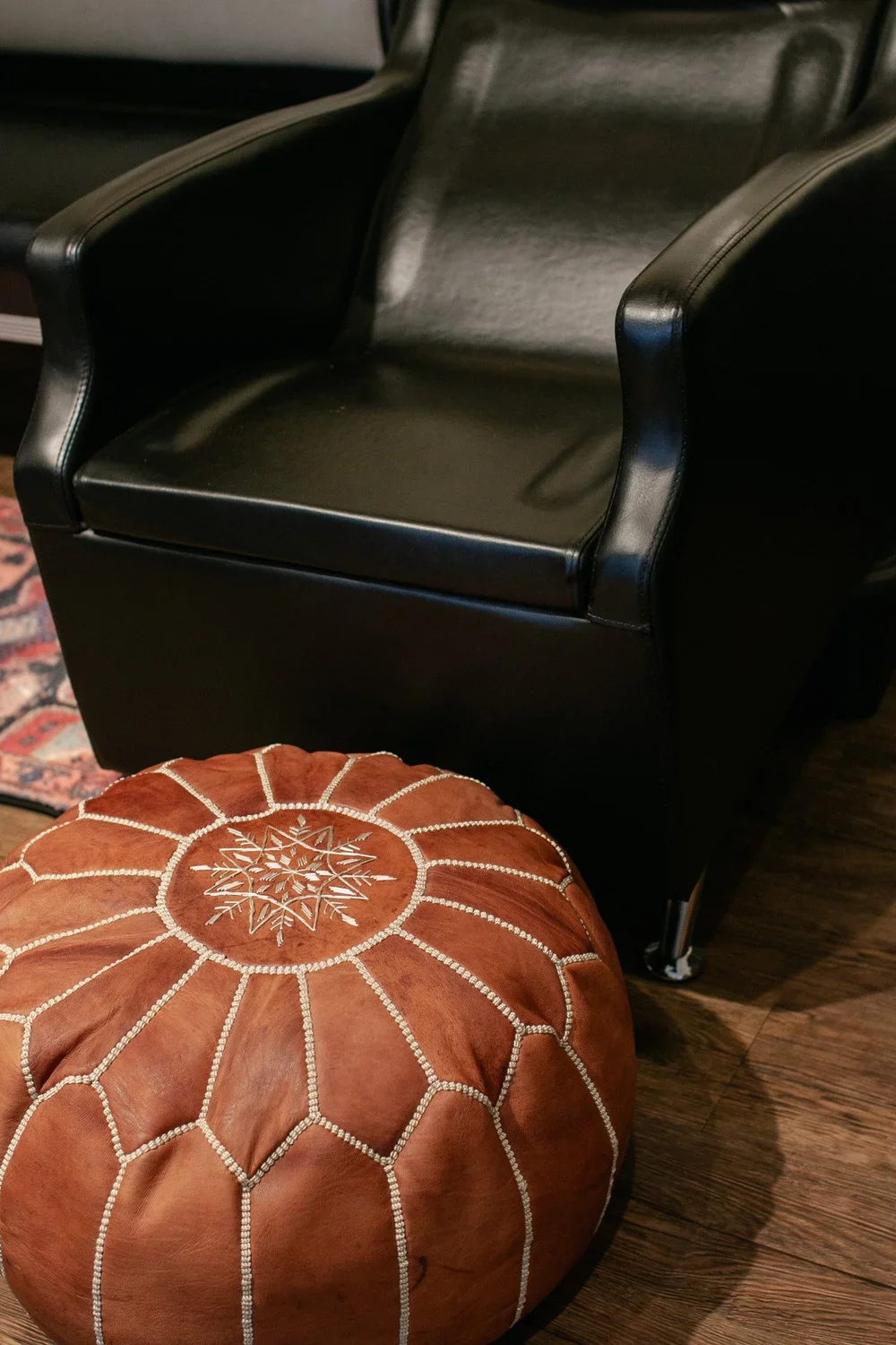 A black leather armchair next to a round, orange-brown leather pouf with white embroidered geometric design, on a wooden floor.