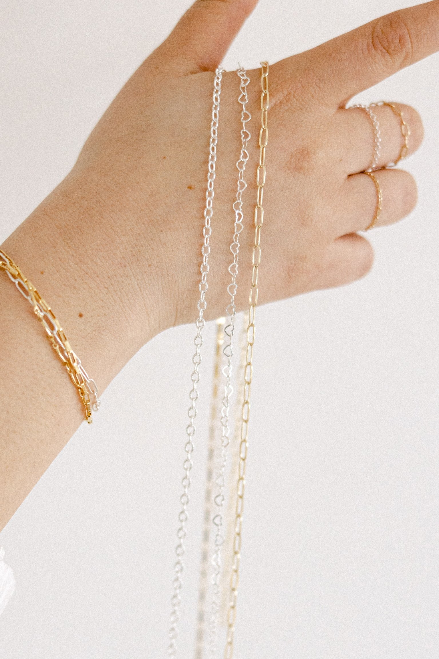 Close-up of a hand with multiple silver and gold chain necklaces and rings, against a white background.