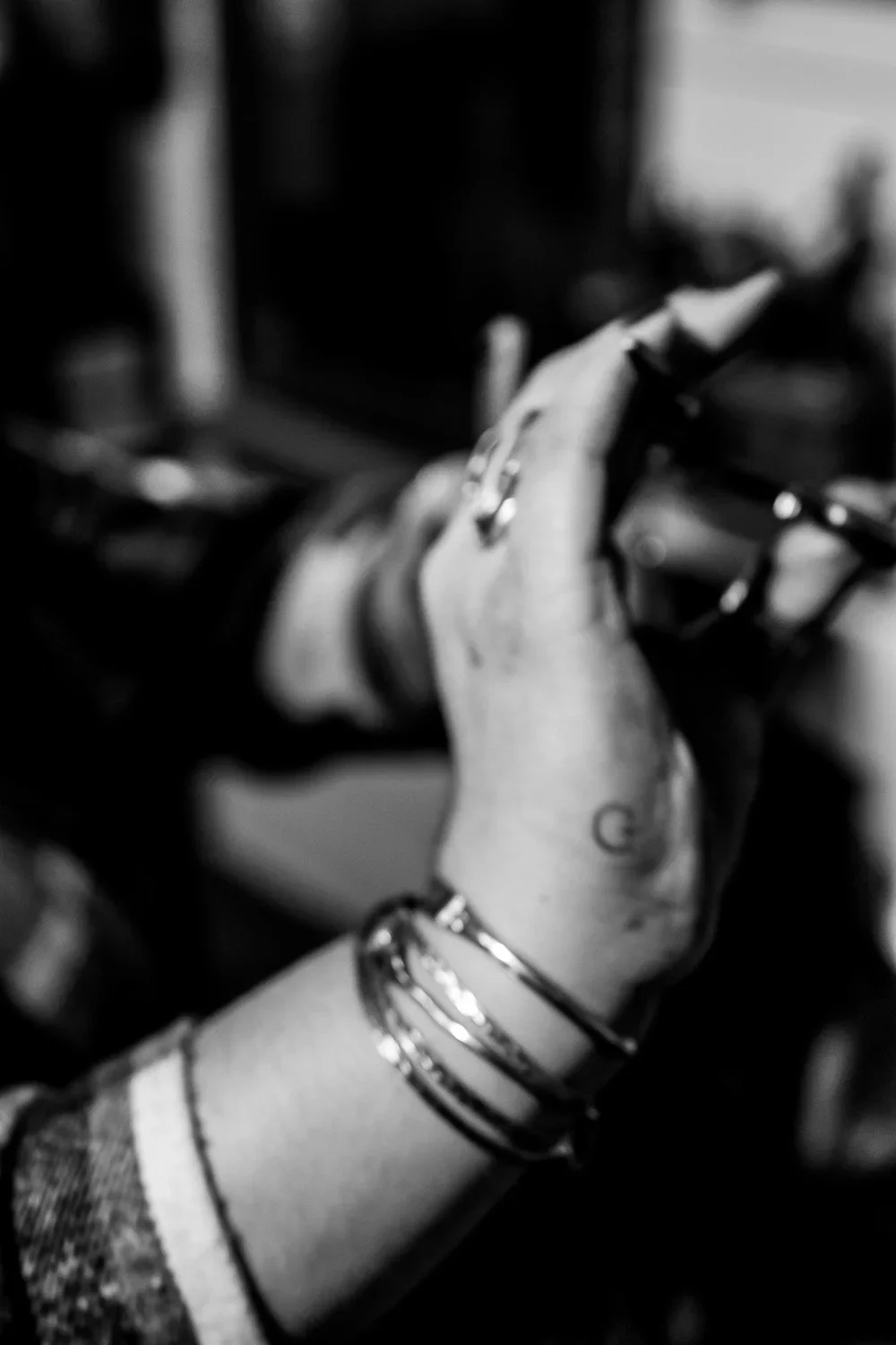 Black and white close-up of a person’s hand holding an object, with multiple bracelets on their wrist and a tattoo visible on their arm.