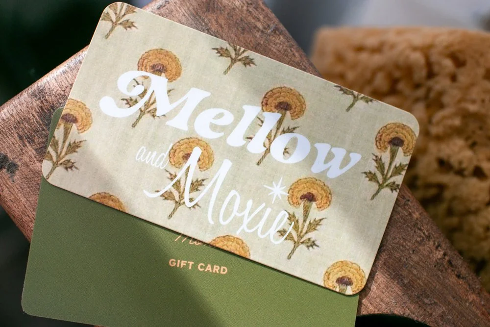 Close-up of a gift card featuring a floral design with yellow flowers and green leaves, with the text 'Mellow and Mouve' on it, placed on a wooden surface.