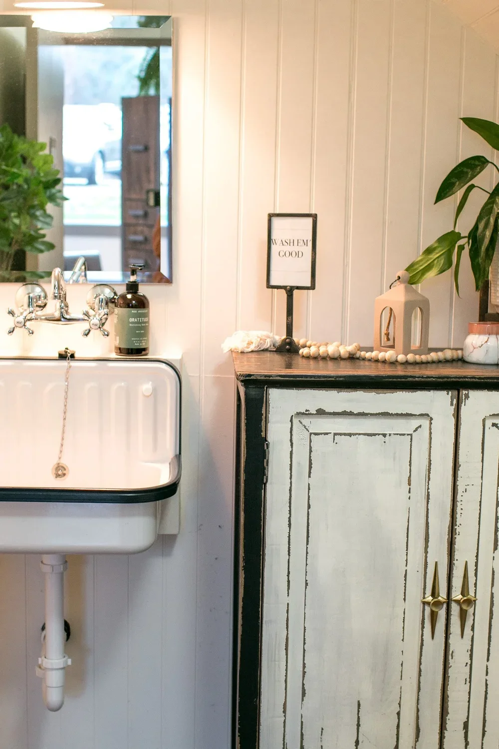 Interior of a vintage-style room with a white ceramic sink, a mirror, a bottle of soap, and a distressed wooden cabinet decorated with a small sign reading "Wash Em Good" and a white decorative lantern, alongside a green plant.