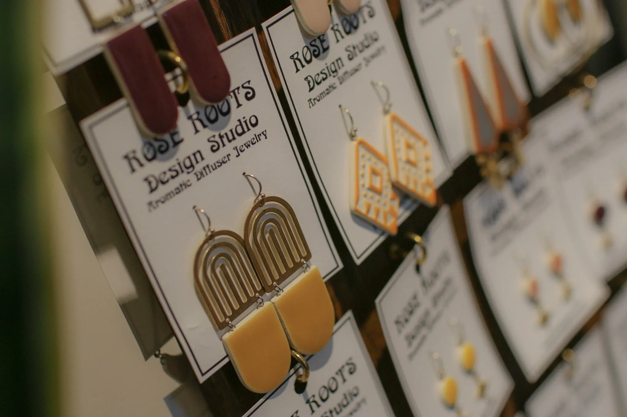 Earrings on display in a jewelry shop, featuring various geometric designs including rainbow, house shape, and abstract styles, labeled as 'Rose Roots Design Studio.'