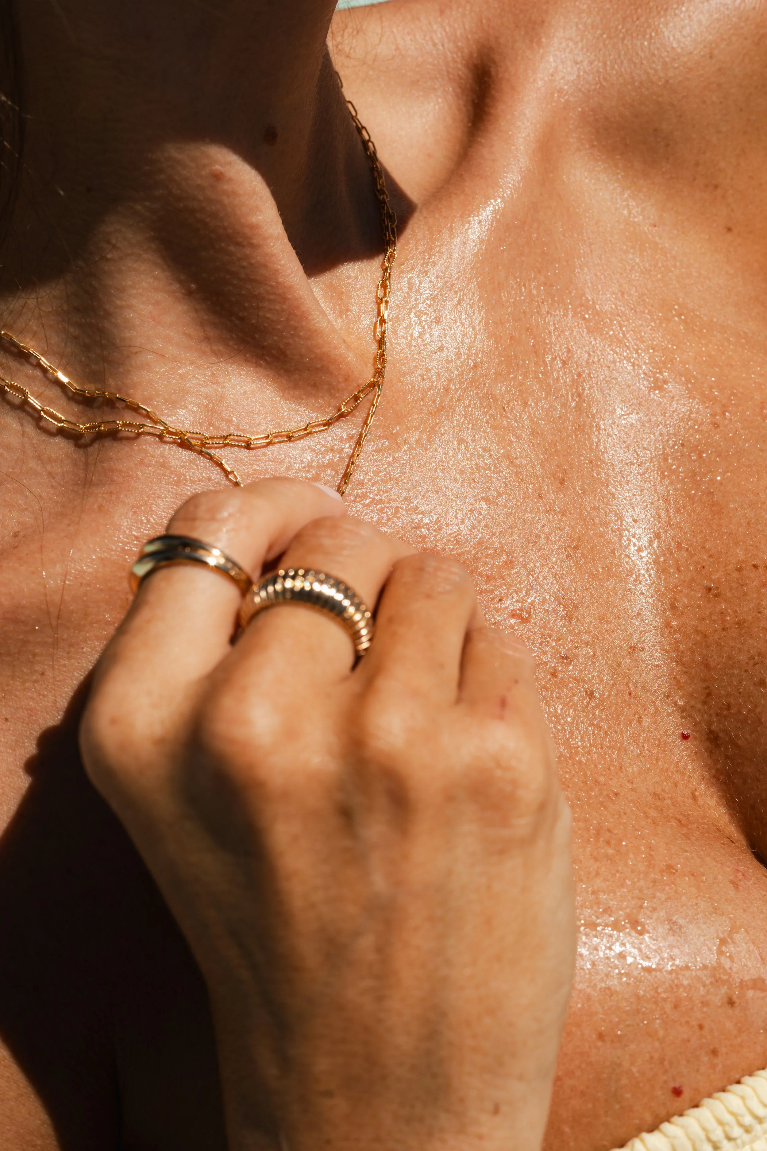 A close-up of a person's neck and hand, showing gold jewelry including layered necklaces and rings, with light reflecting off their skin.