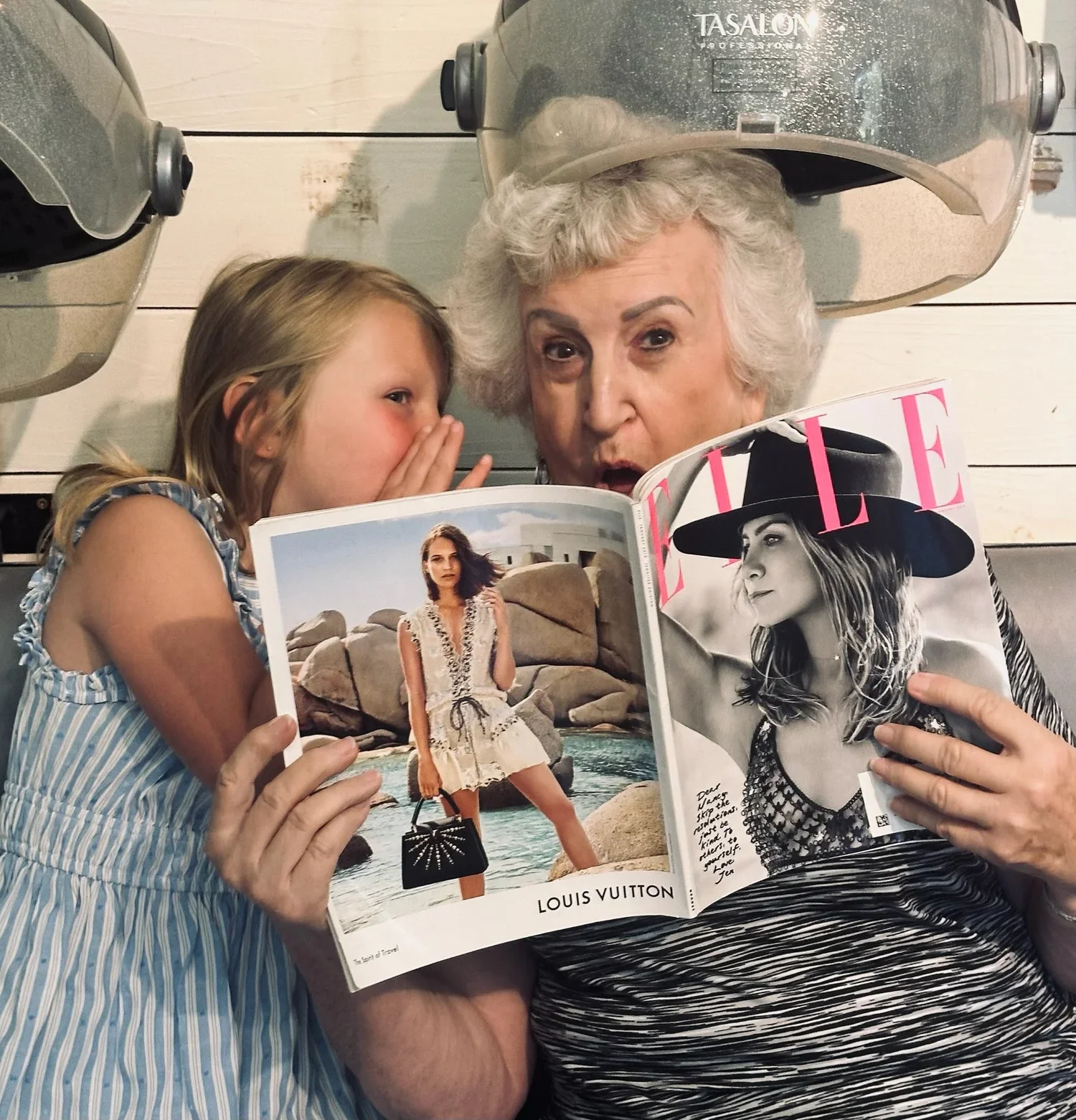 An elderly woman and a young girl are lying under hair dryers in a salon, reading a magazine together. The woman is holding the magazine open, showing a fashion spread, while the girl whispers into her ear.
