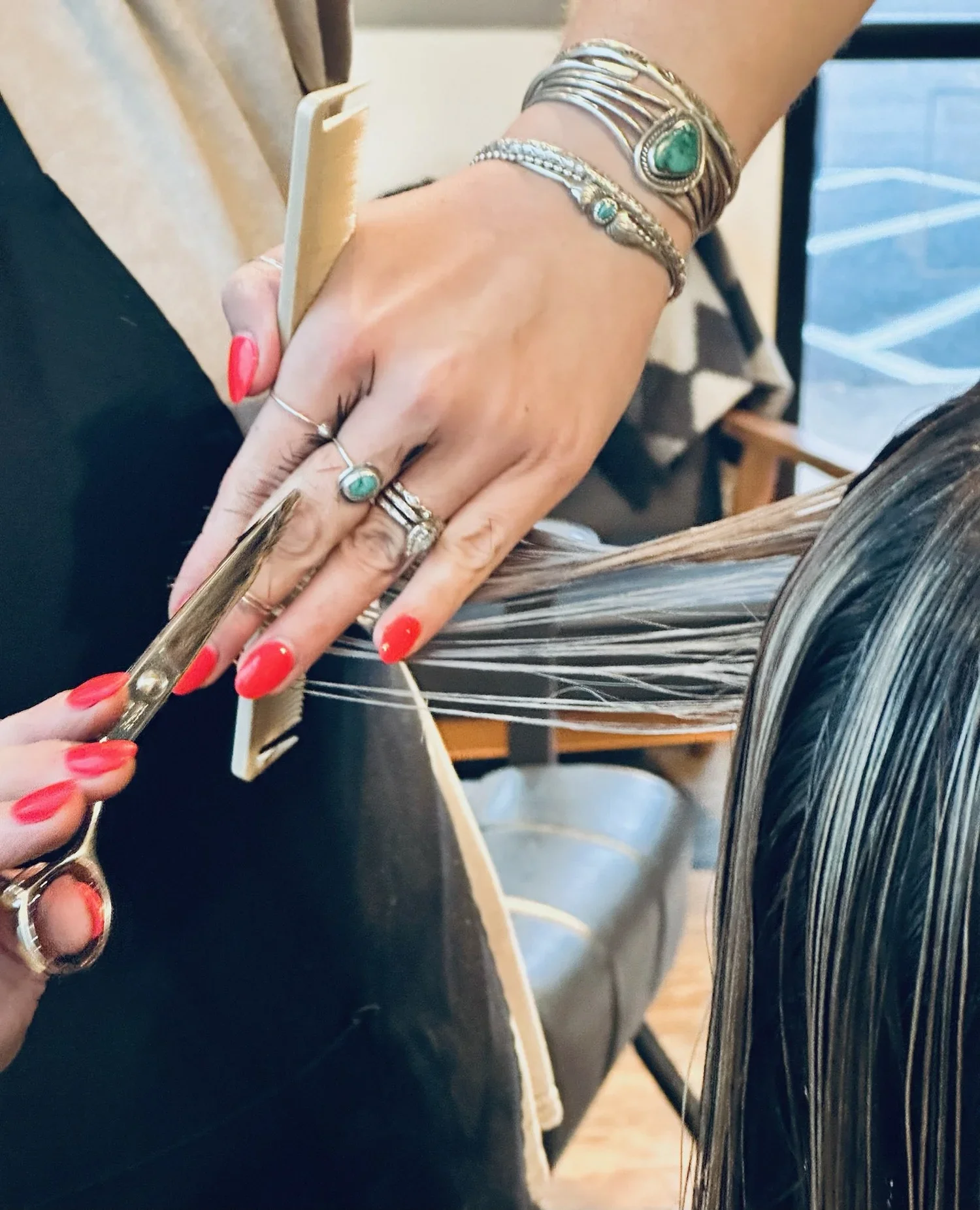 A hairstylist cutting a client's light brown hair with scissors, while holding a section with a comb. The stylist is wearing rings, bracelets, and has painted nails.
