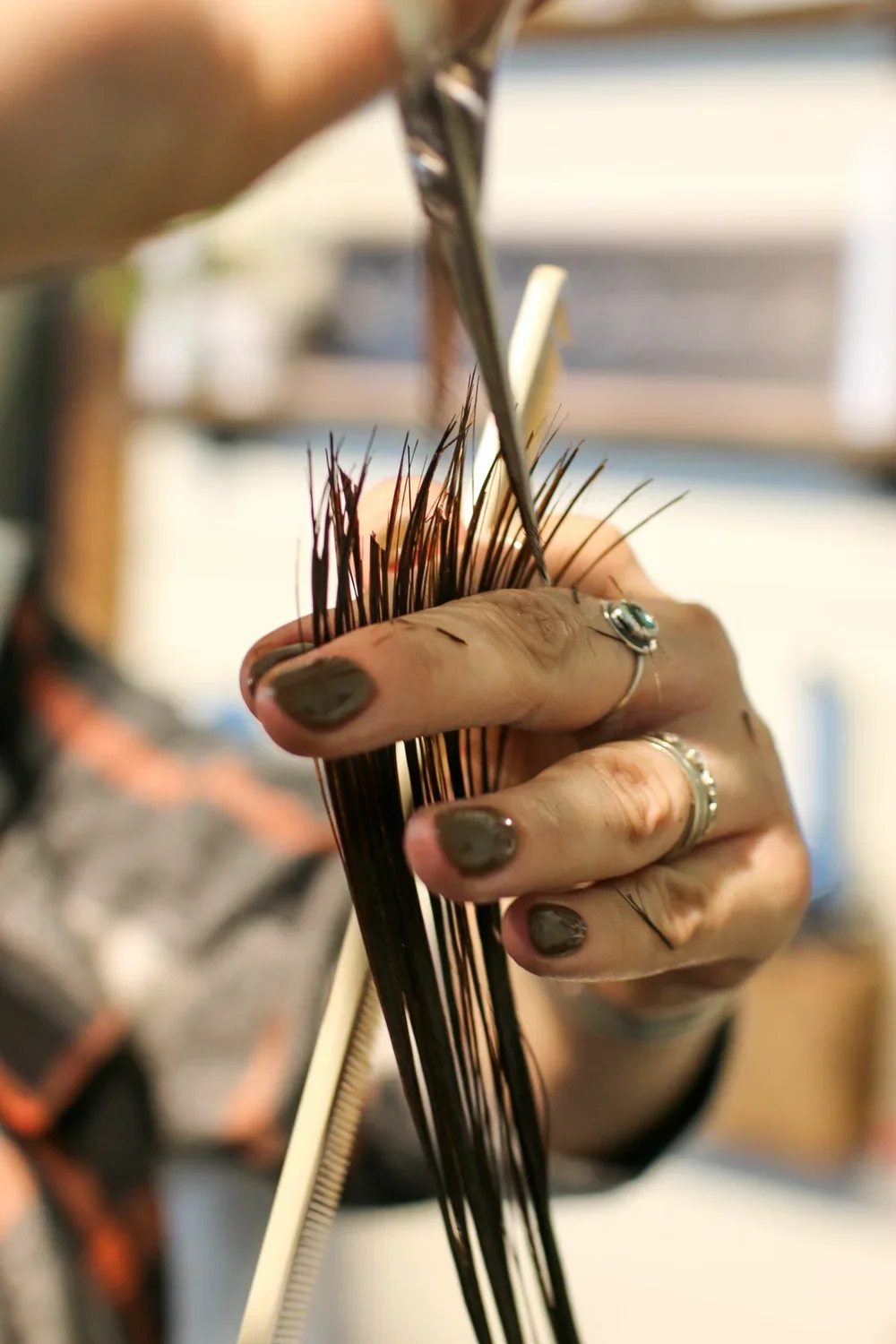 Close-up of a person's hand with rings, weaving or handling dark brown or black hair extensions or strands with a metal tool in a well-lit indoor setting.