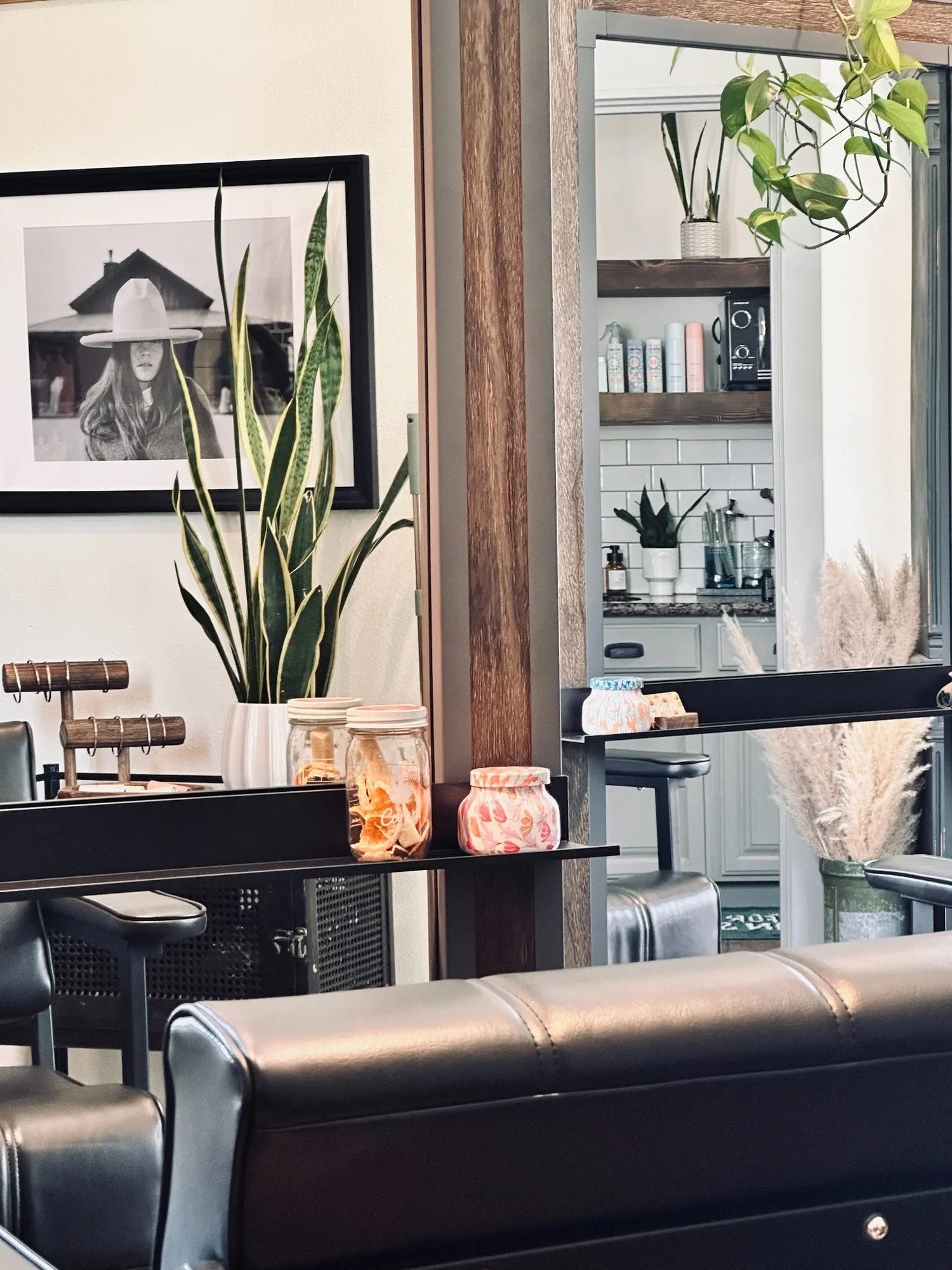 Interior view of a cozy cafe showing black leather seating, decorative plants, a framed black and white photograph of a woman, and a kitchenette in the background with shelves and containers.