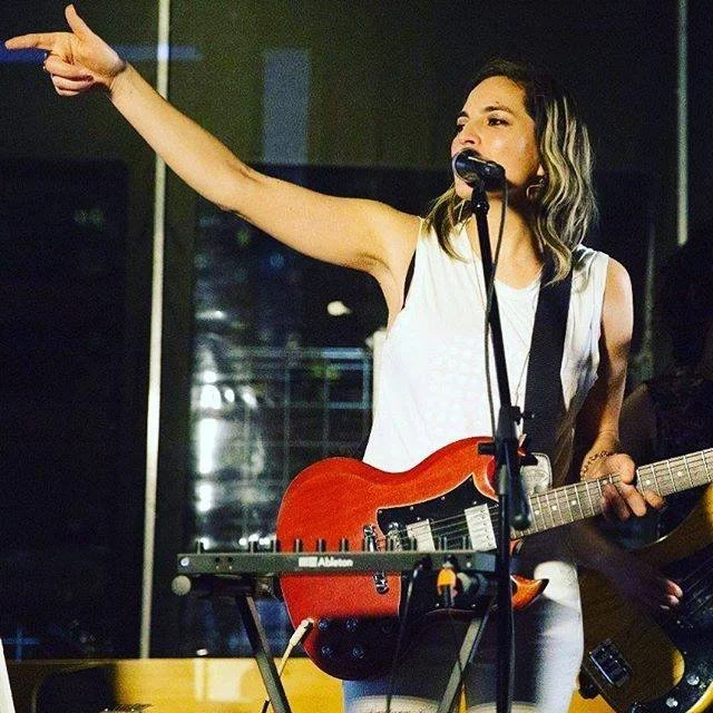 A woman with shoulder-length hair, wearing a sleeveless white top, is holding an electric guitar and singing into a microphone. She is extending her right arm outward with her index finger pointing, possibly performing on stage.