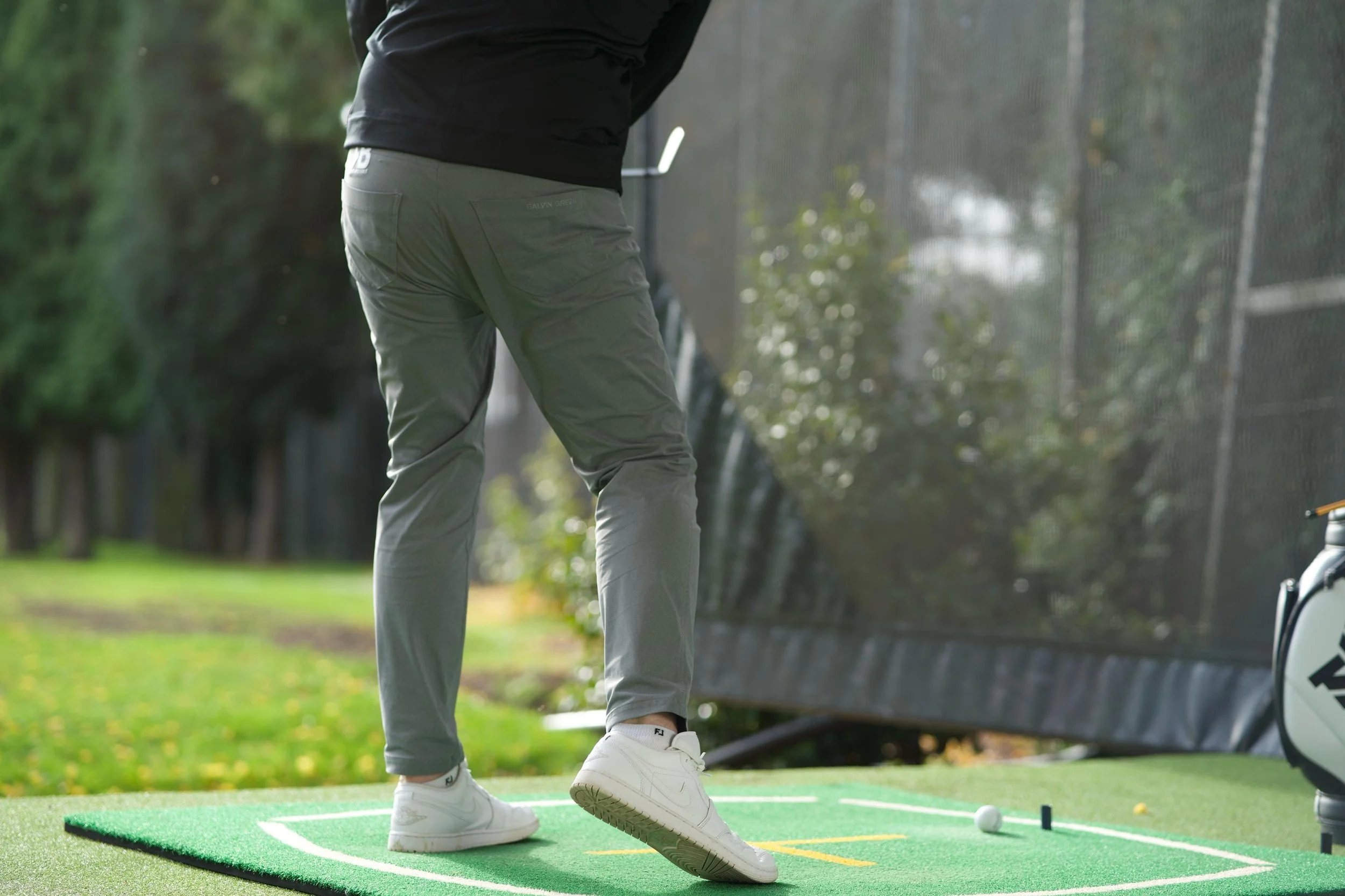 Person practicing golf on a driving range, focusing on their lower body and golf club, with a golf bag nearby.