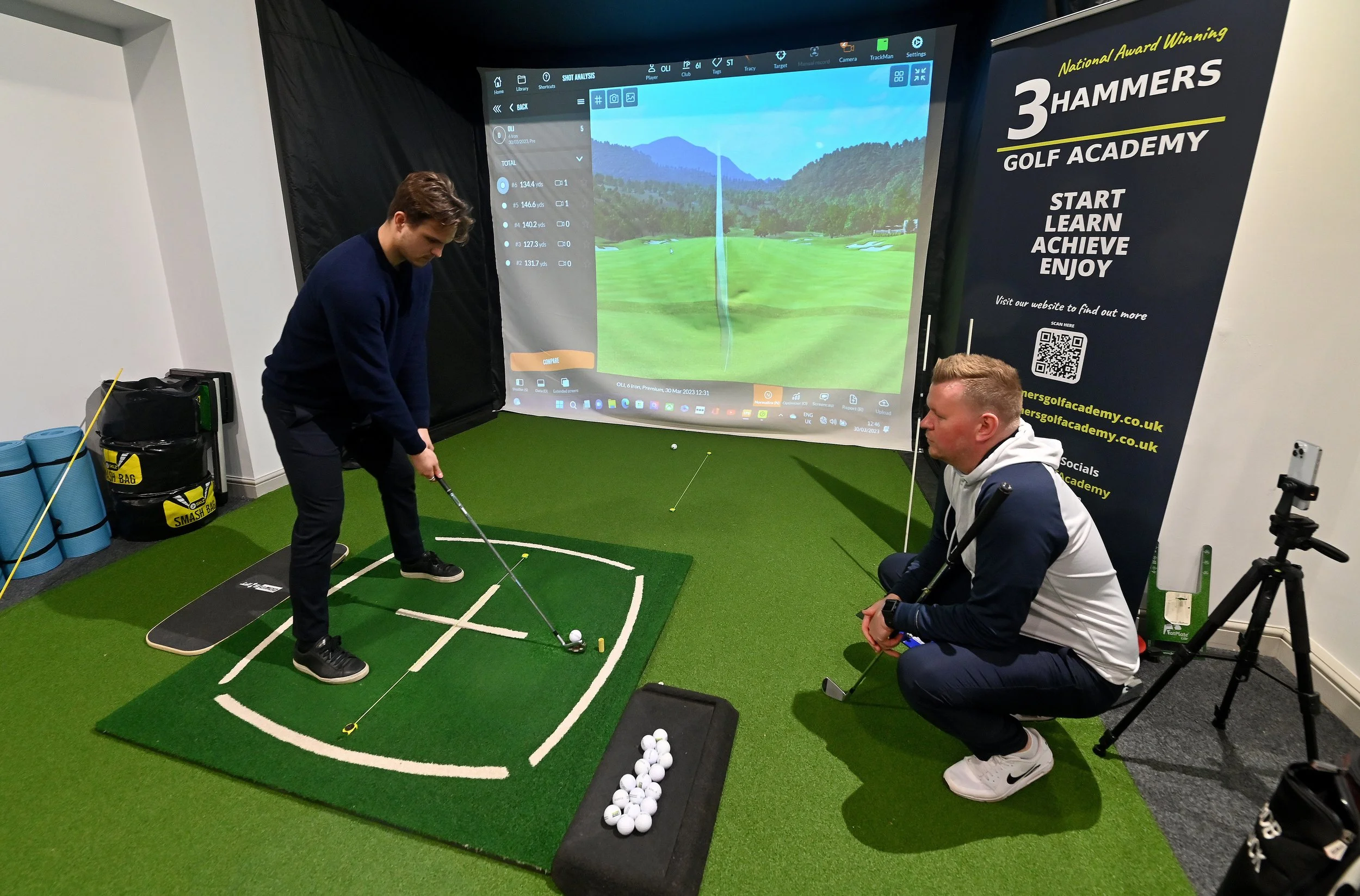 Two men in a golf simulation room; one preparing to hit a golf ball on a small indoor grass mat, with a large screen displaying a virtual golf course in front of them. The room has a sign that reads '3 Hammer Golf Academy' with the words 'Start, Lear
