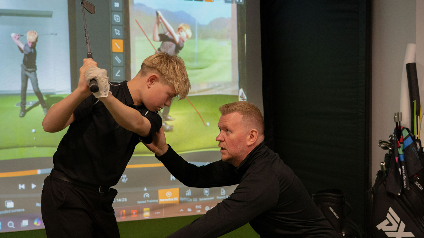 A golf instructor assists a young male golfer with his swing technique, inside an indoor golf training facility with a virtual golf simulator in the background.