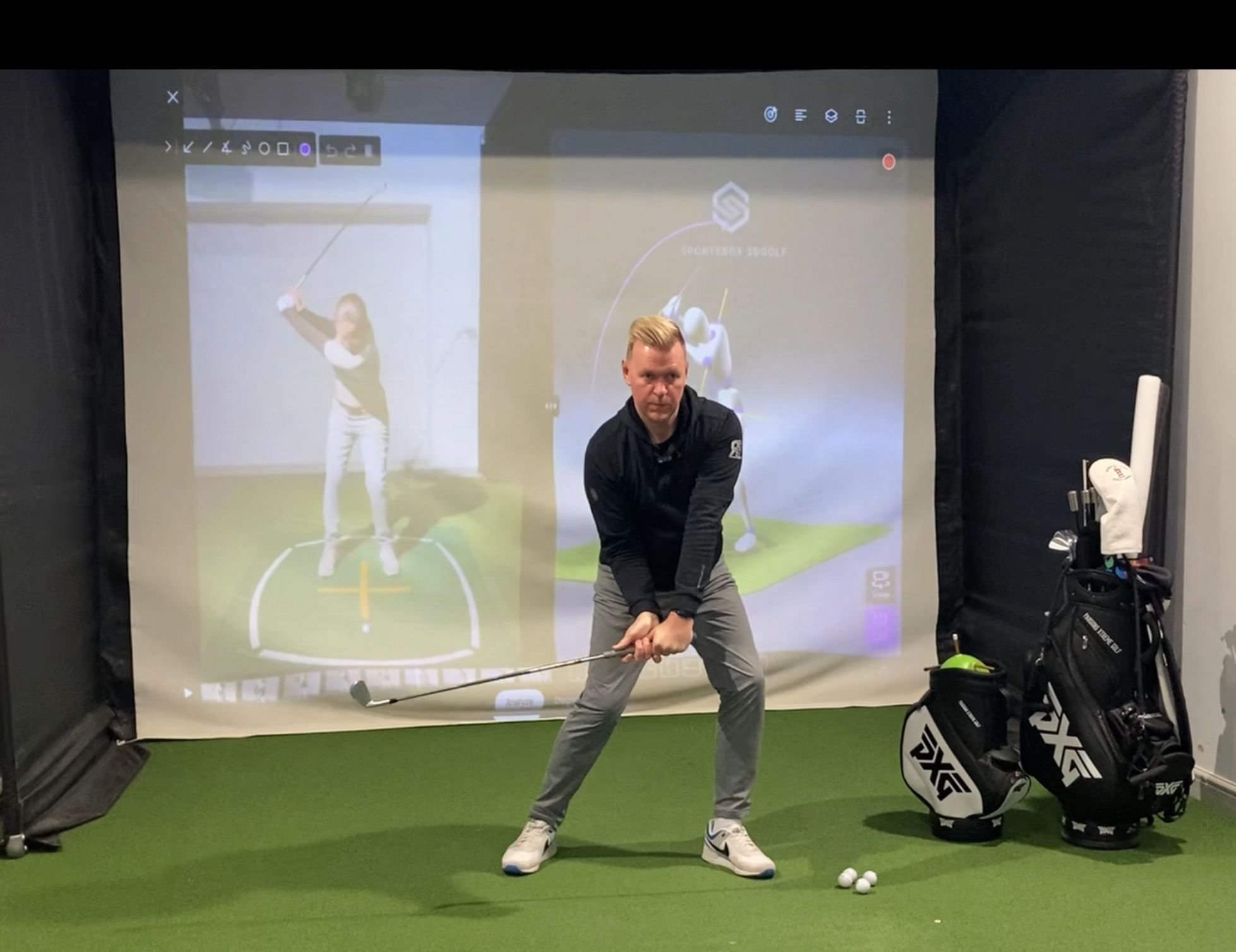 Man practicing golf swing in indoor golf simulator with golf bags and balls on green turf.