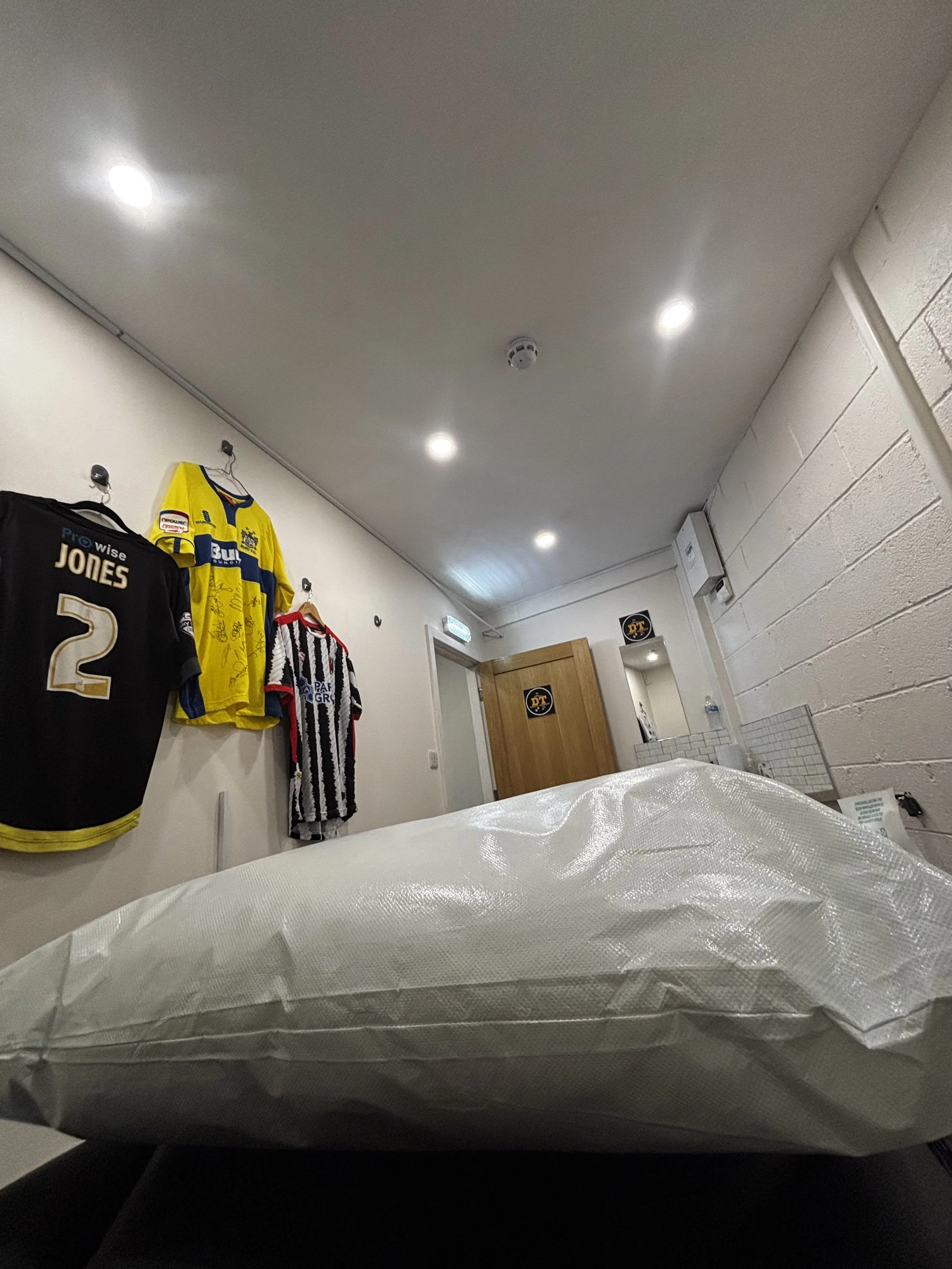 Interior of a room with white walls and ceiling, illuminated by several ceiling lights. Three sports jerseys are hanging on the wall: a black one with the name 'Jones' and the number 2, a yellow and blue one, and a black and white striped one. A bed or examination table with a white cover is in the foreground.