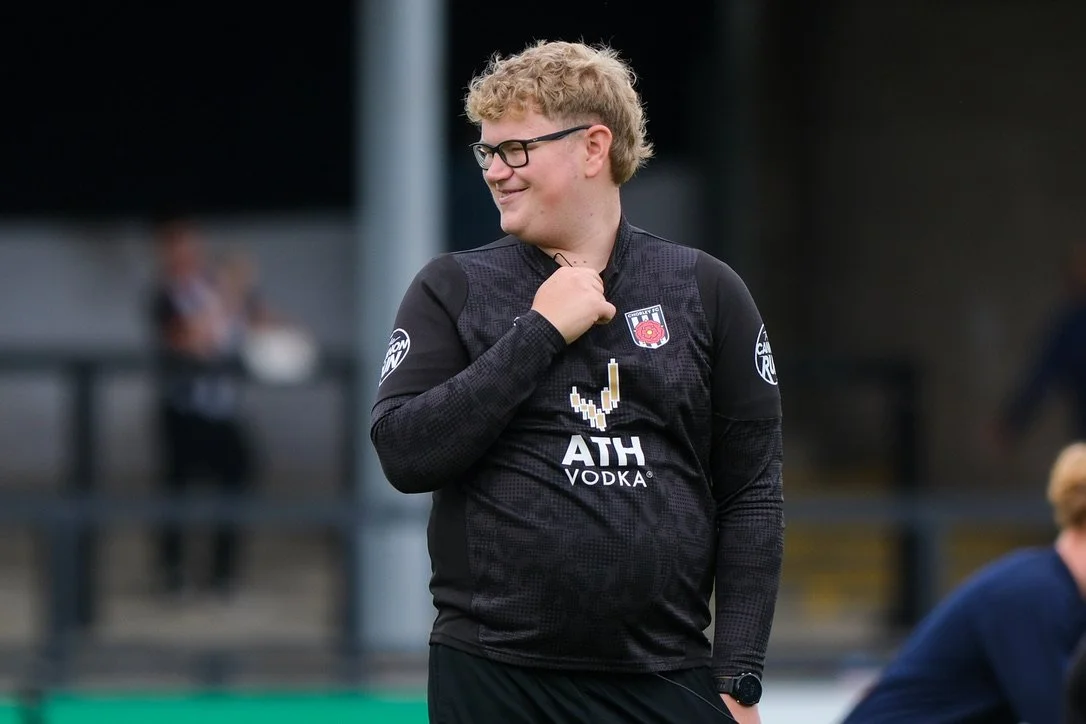 A young man with curly blonde hair, glasses, and a black sports jersey smiling and adjusting his collar during a sports event.