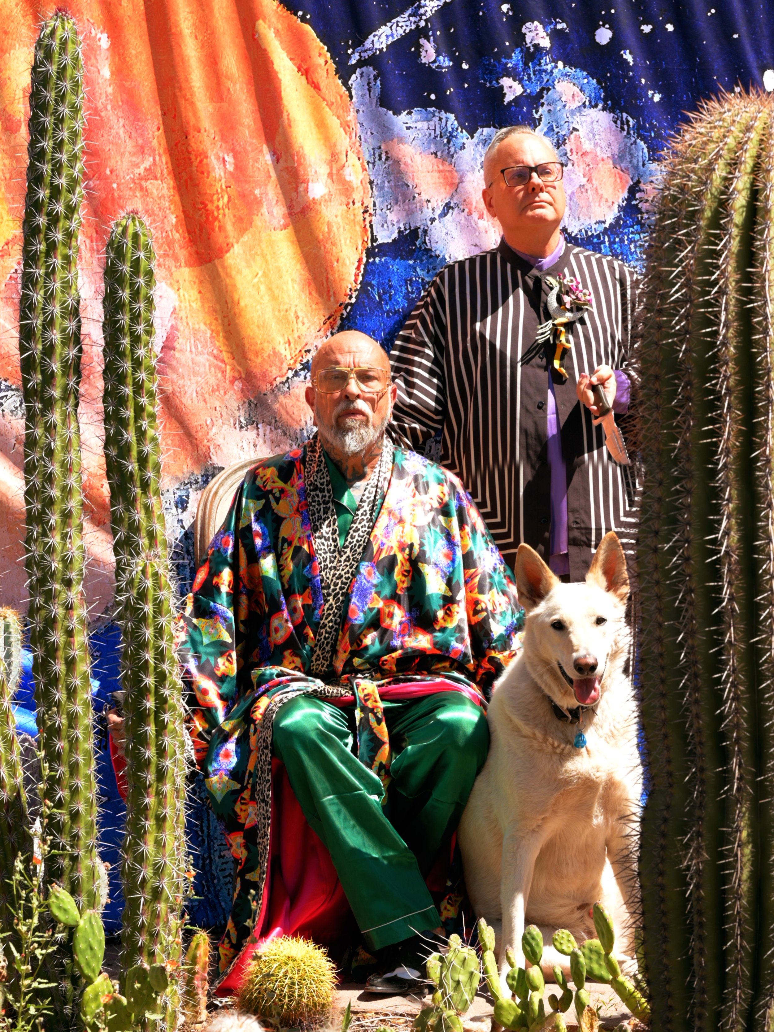 Mark Mitchell, Kurt B Reighley, and rescue dog Ghost jin the midst of their cactus garden before a backdrop from a painting by Mark Mitchell, Tucson AZ