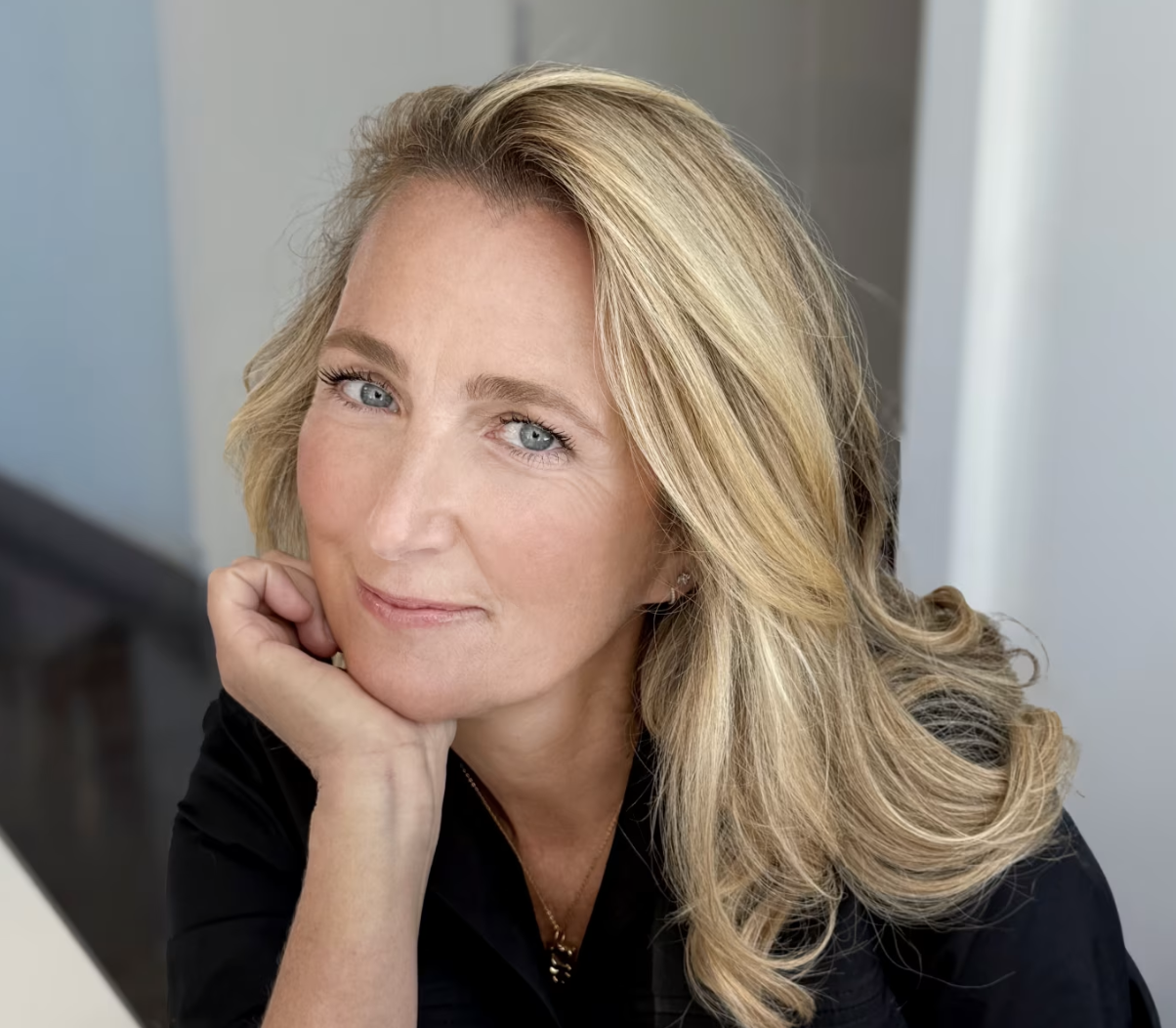 Close-up portrait of a middle-aged woman with blonde hair, blue eyes, and light makeup, resting her chin on her hand and smiling slightly.
