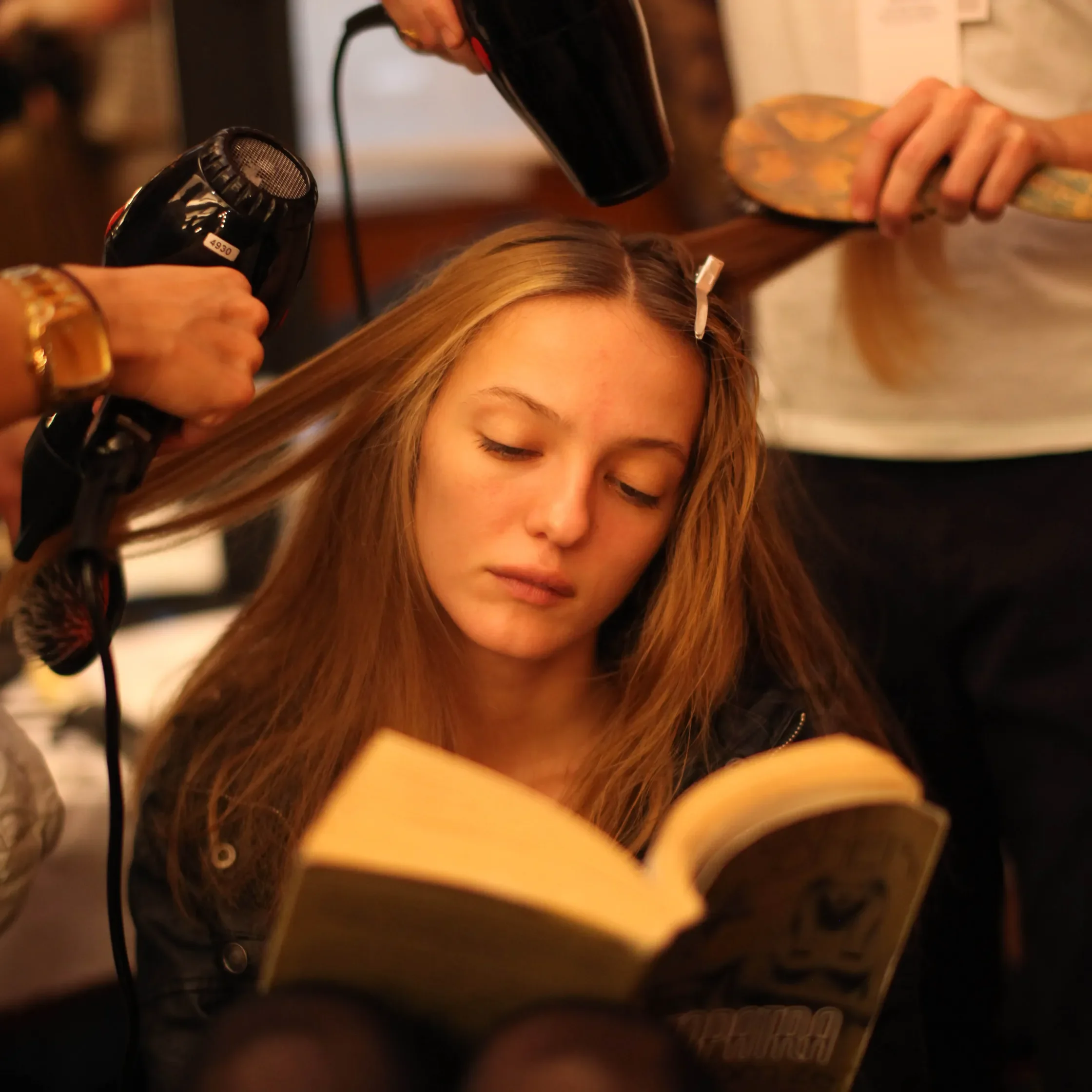 Person styling long blonde hair with a hairdryer and a paddle brush at a salon.