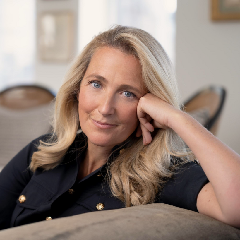 A woman with blonde hair and blue eyes resting her head on her hand and smiling at the camera in a well-lit indoor setting.