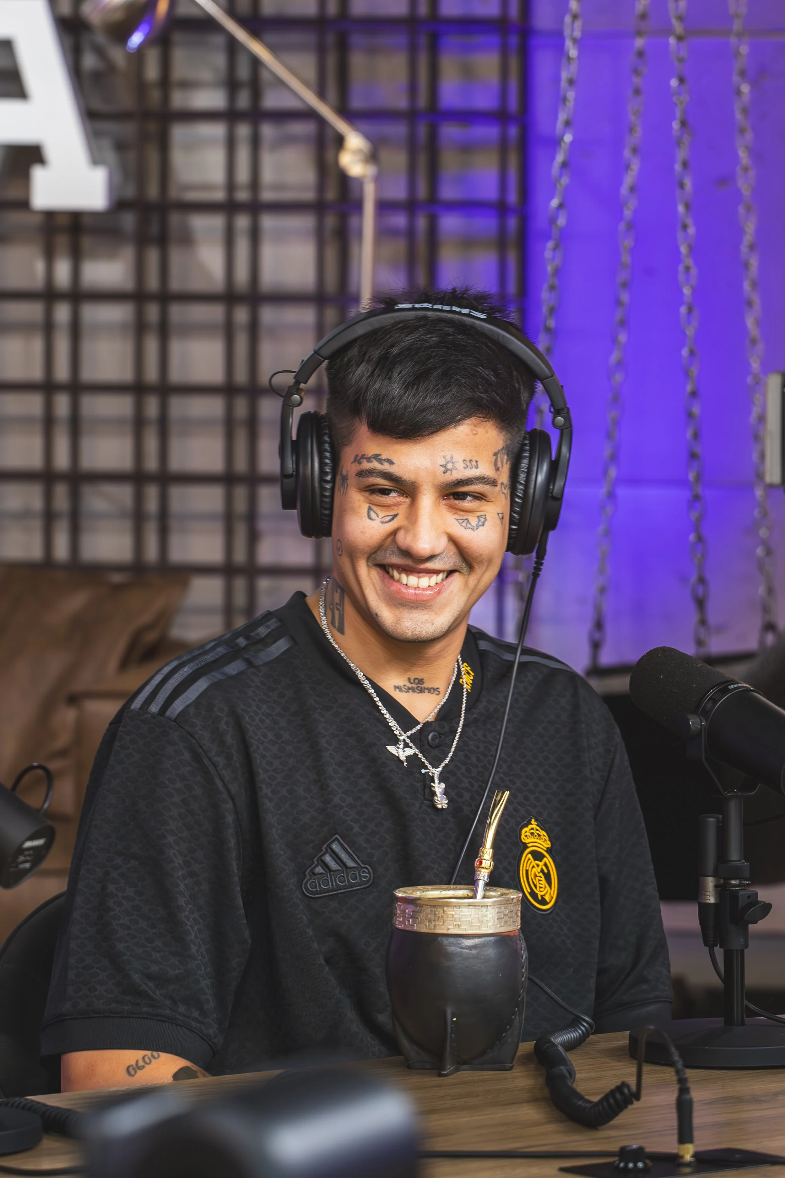 Joven con tatuajes en la cara, usando audífonos grandes y camiseta de fútbol, sonriendo frente a un micrófono en un estudio de grabación.