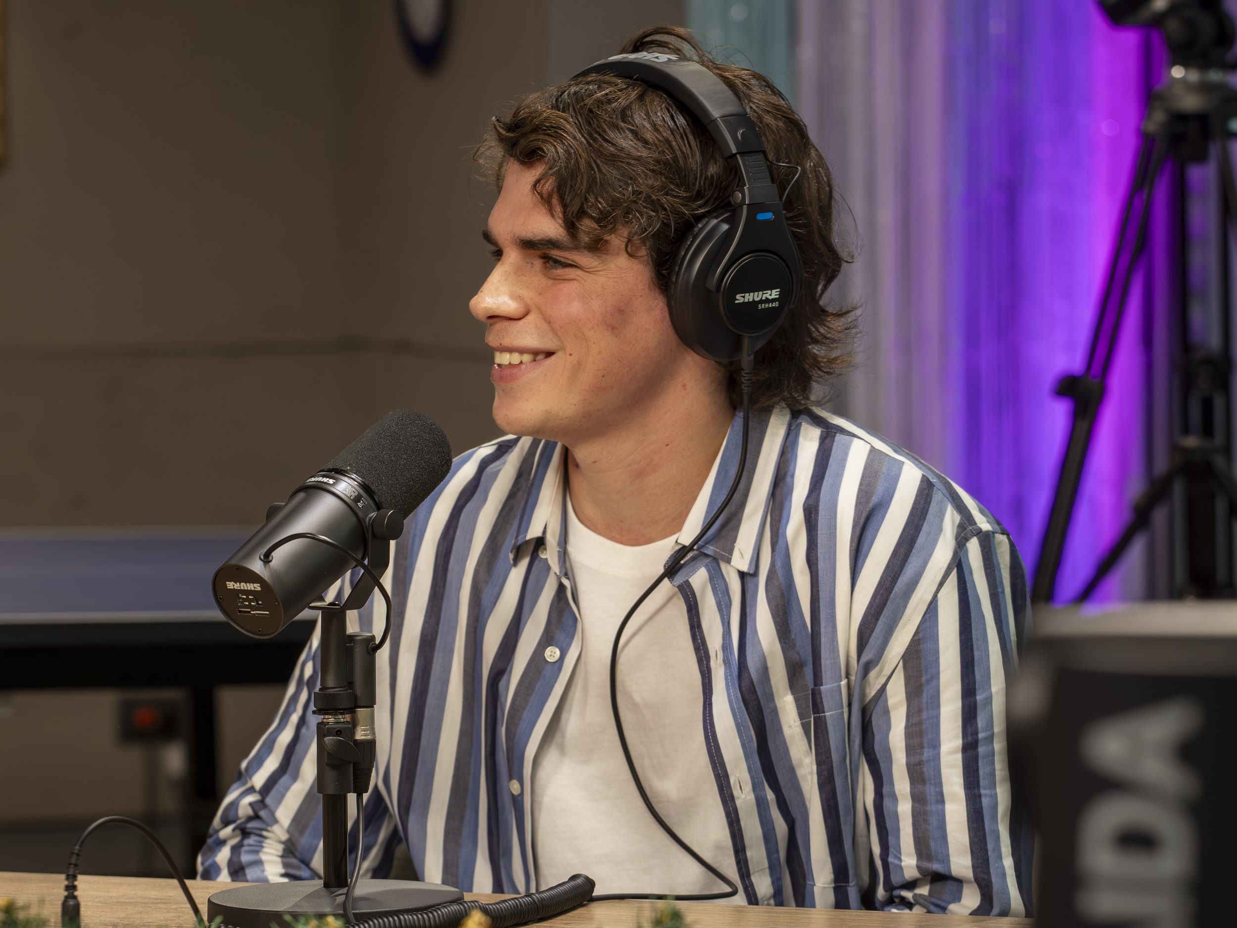 Joven hombre con cabello rizado usando audífonos y camiseta a rayas, sonriendo en un estudio de grabación con micrófono frente a él.
