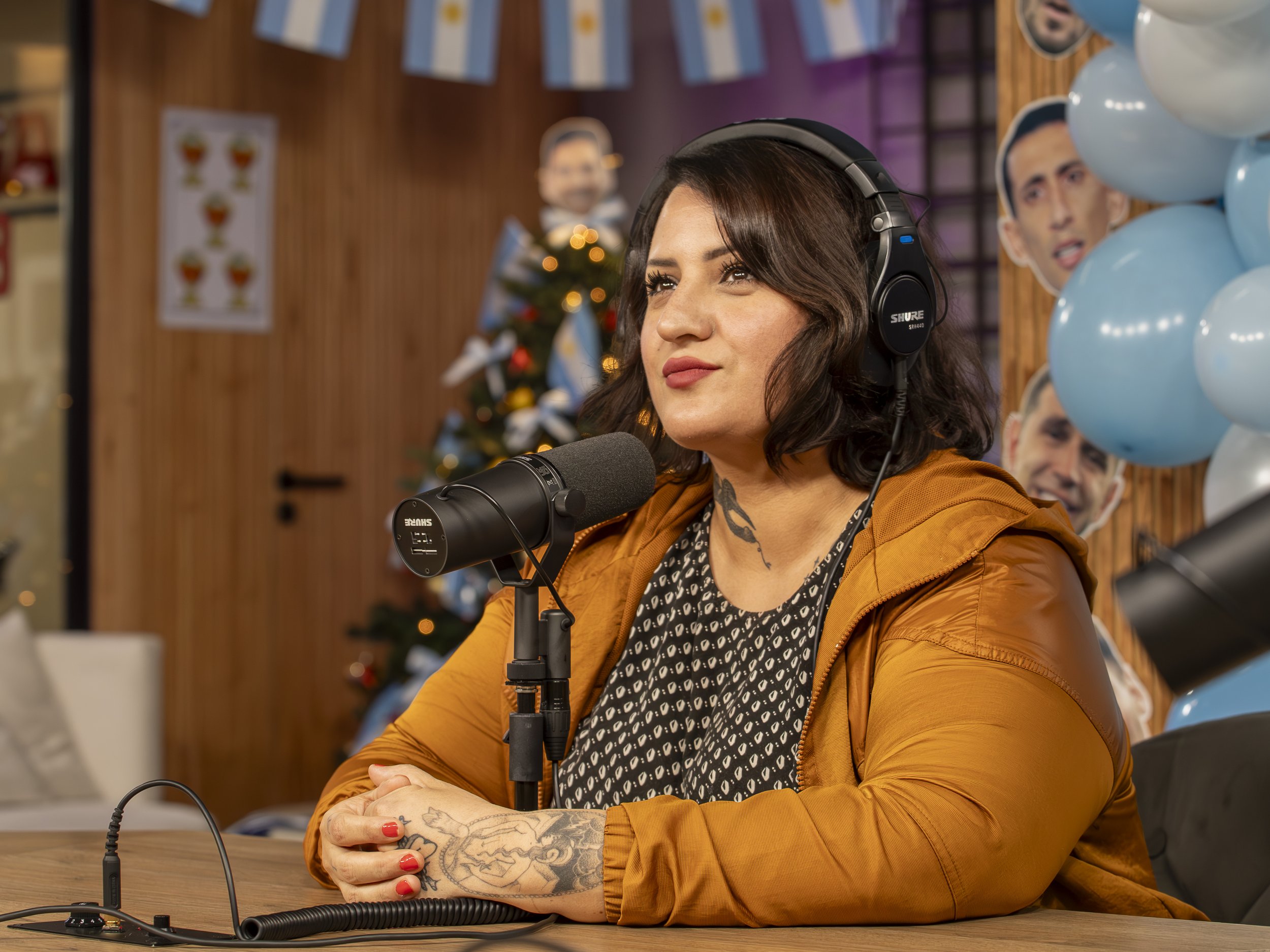 Una mujer con auriculares y tatuajes, hablando en un micrófono en un estudio decorado con globos y un árbol de Navidad.