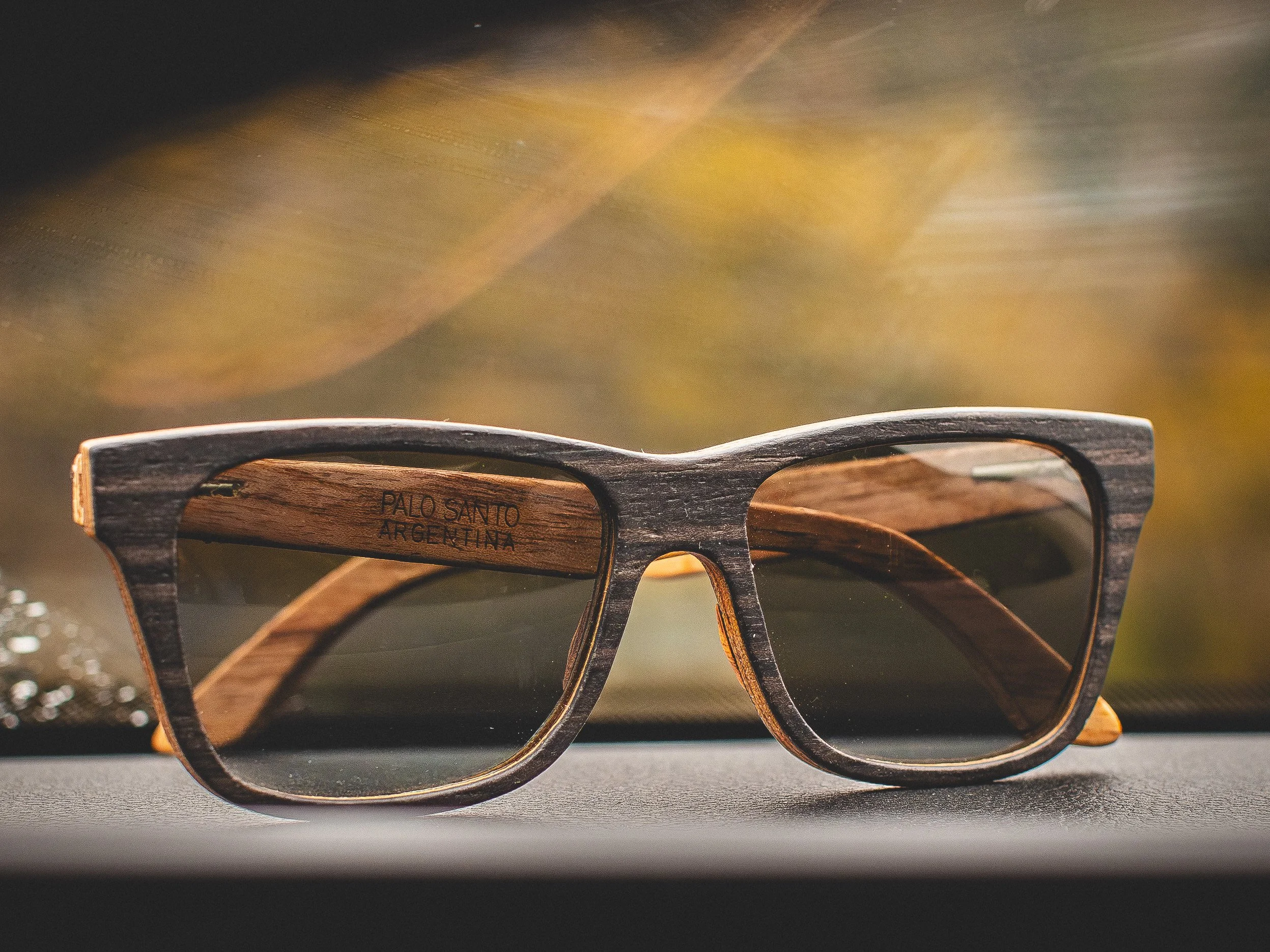Gafas de sol con marco de madera y lentes oscuros, descansando en un tablero con fondo borroso de tonos cálidos.