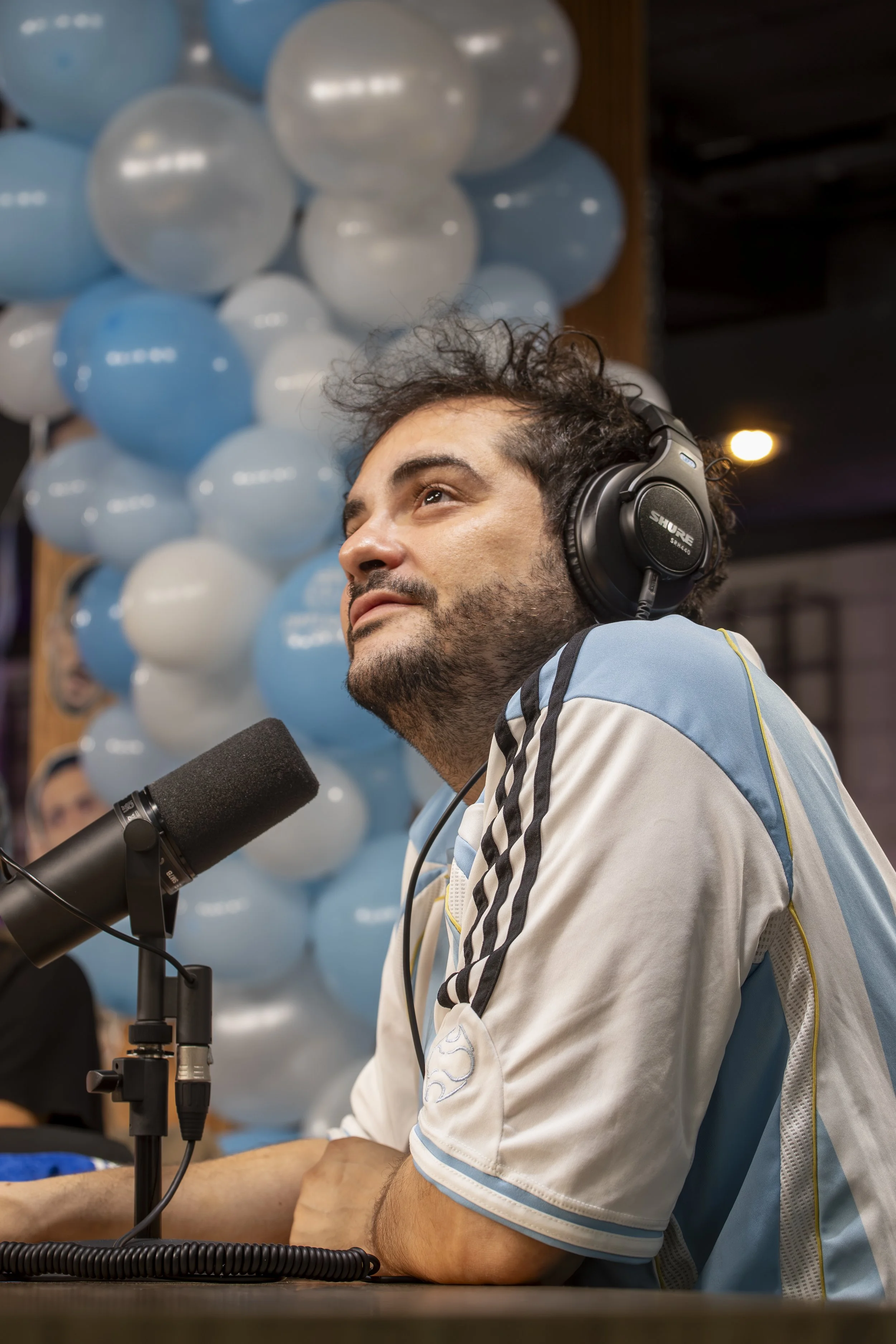 Hombre con auriculares en un estudio de grabación, sentado frente a un micrófono, con globos en el fondo.