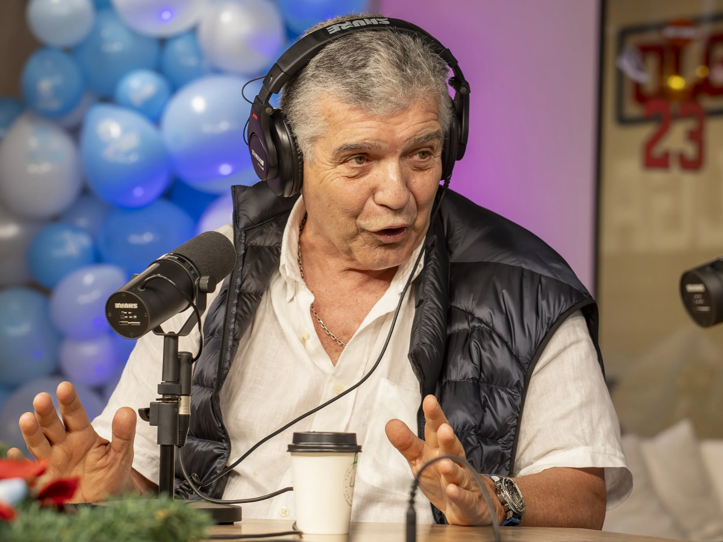 Un hombre mayor con cabello gris, usando auriculares grandes, una camisa blanca y un chaleco negro, hablando en un micrófono en un estudio de grabación o podcast, con globos azules al fondo y un cartel de baloncesto en la pared.